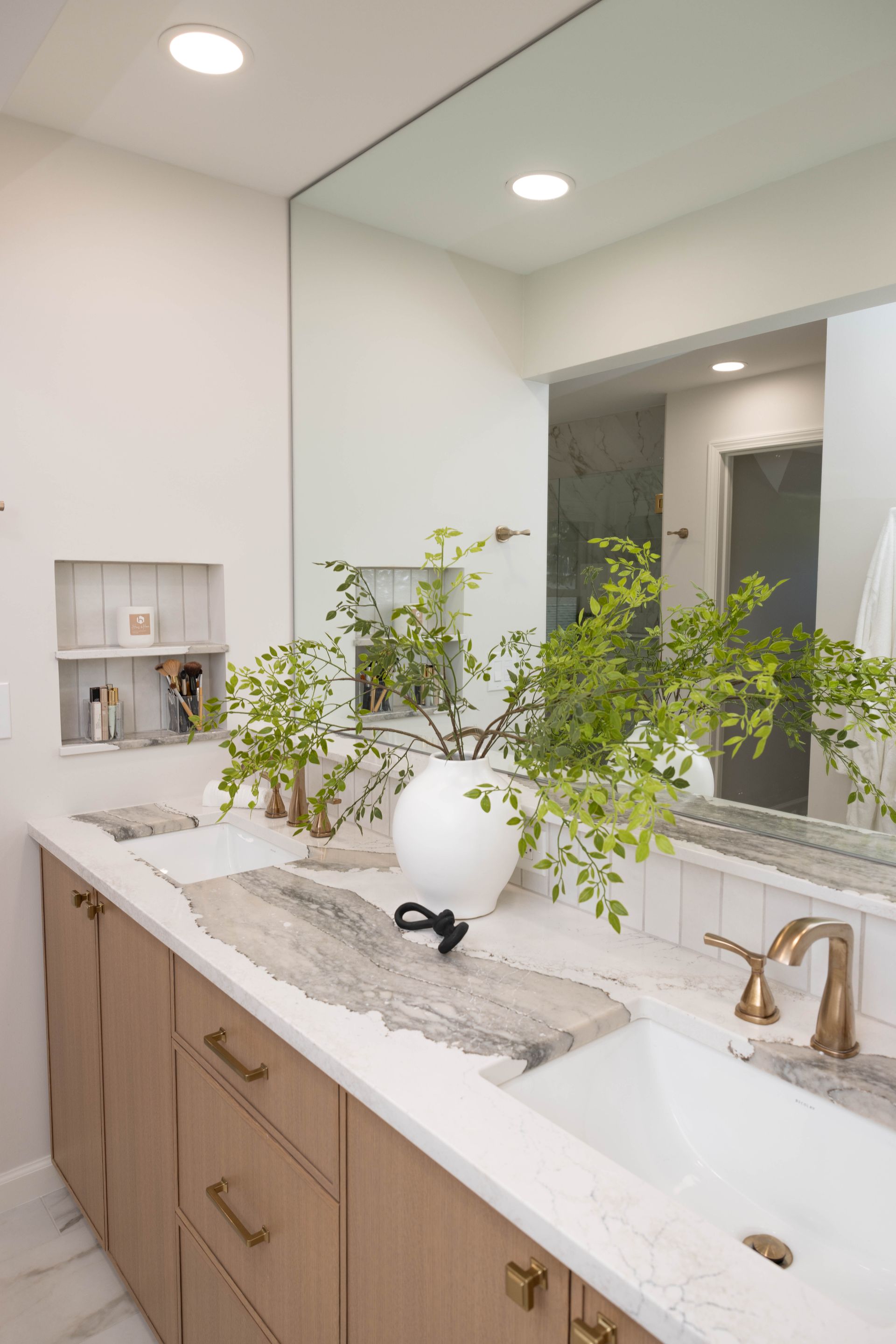 Modern bathroom with light wood cabinets, marble countertop, large mirror, and green plant decor.