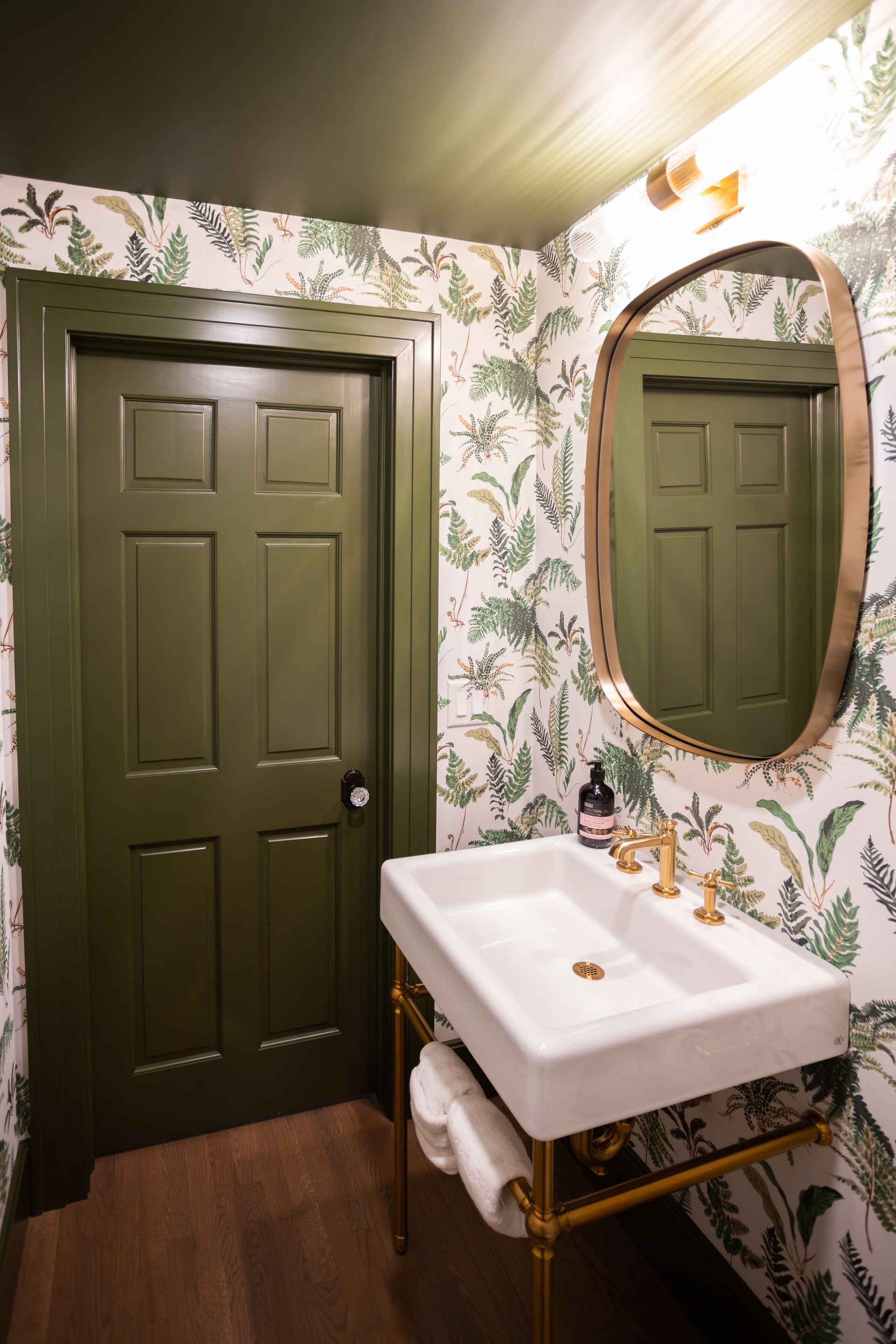 Green powder room with botanical wallpaper, olive-green door, gold fixtures, and oval mirror.
