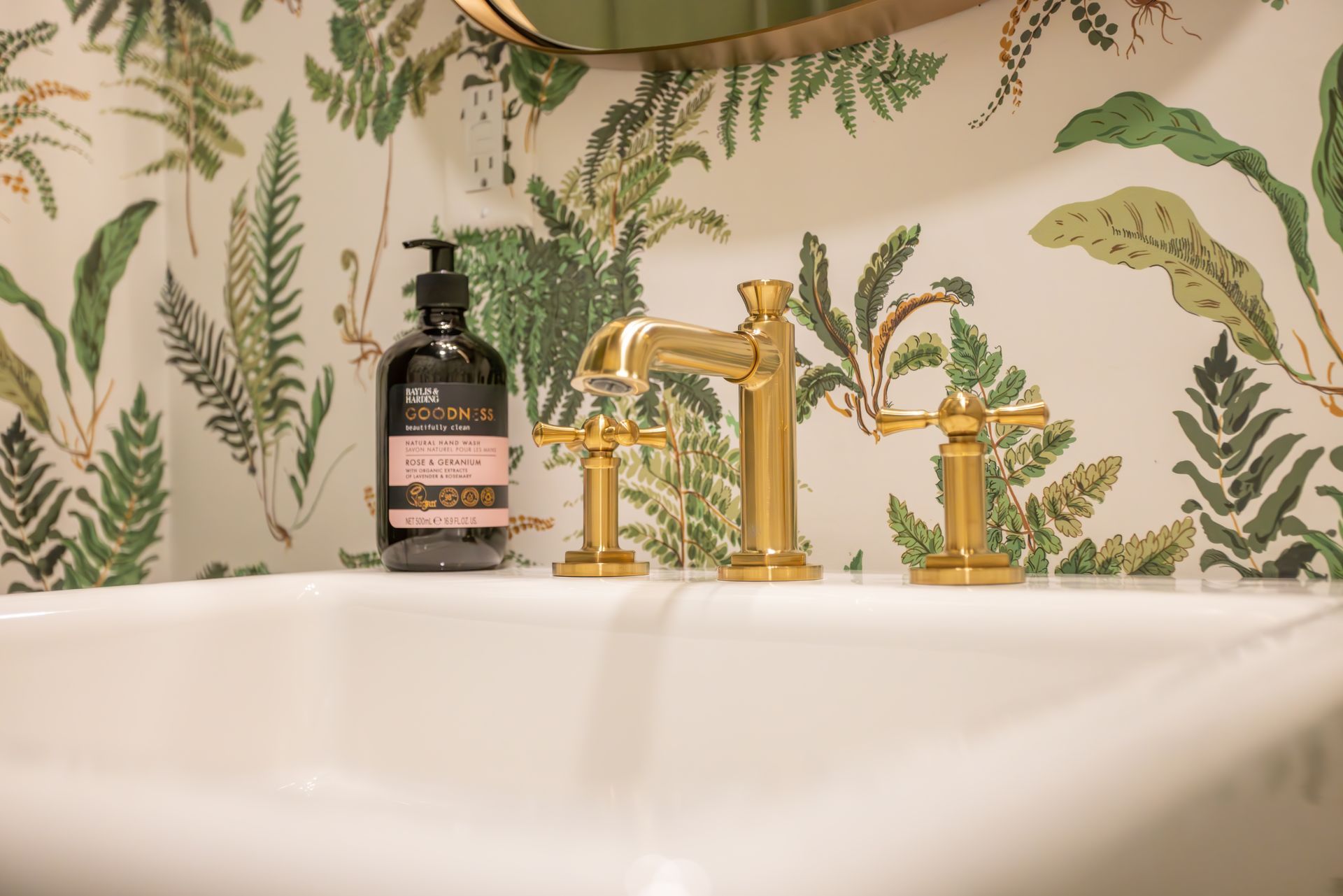 White sink with gold faucet and fern wallpaper, black soap dispenser.