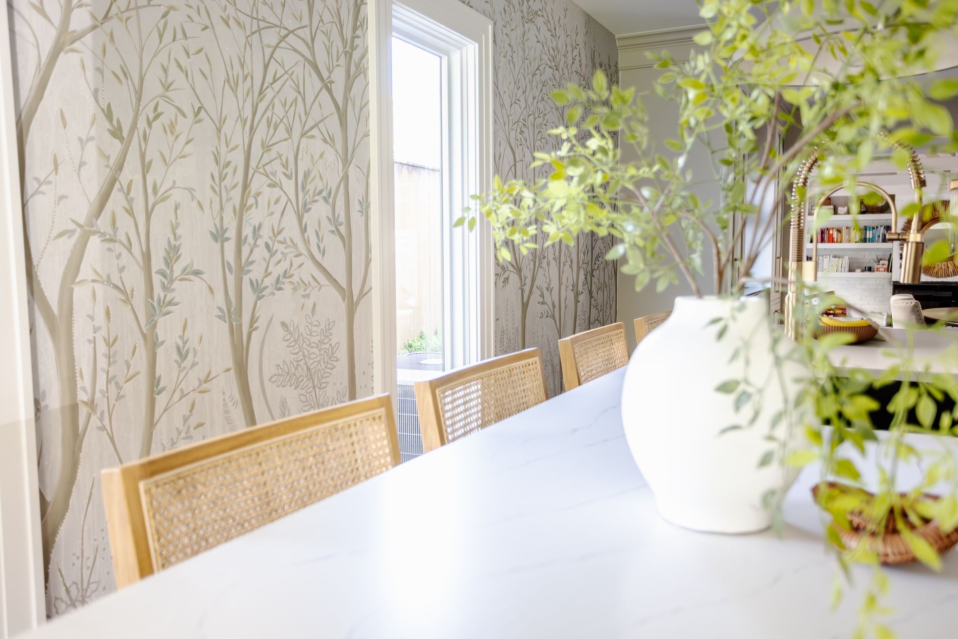 Dining room with white table, rattan chairs, and tree-patterned wallpaper. A vase with greenery sits on the table.