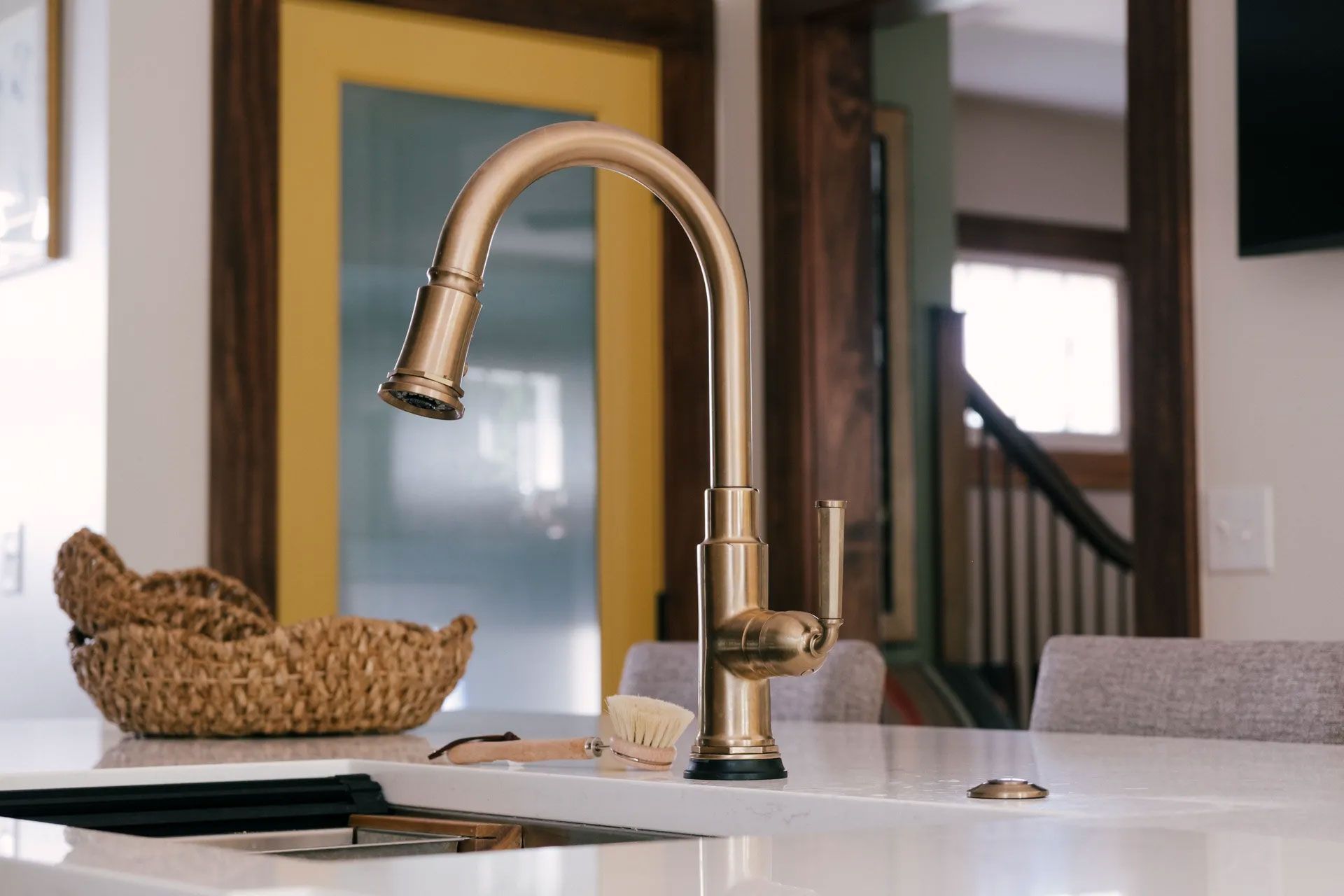 Gold kitchen faucet on white countertop. Basket, brush, and yellow door in background.