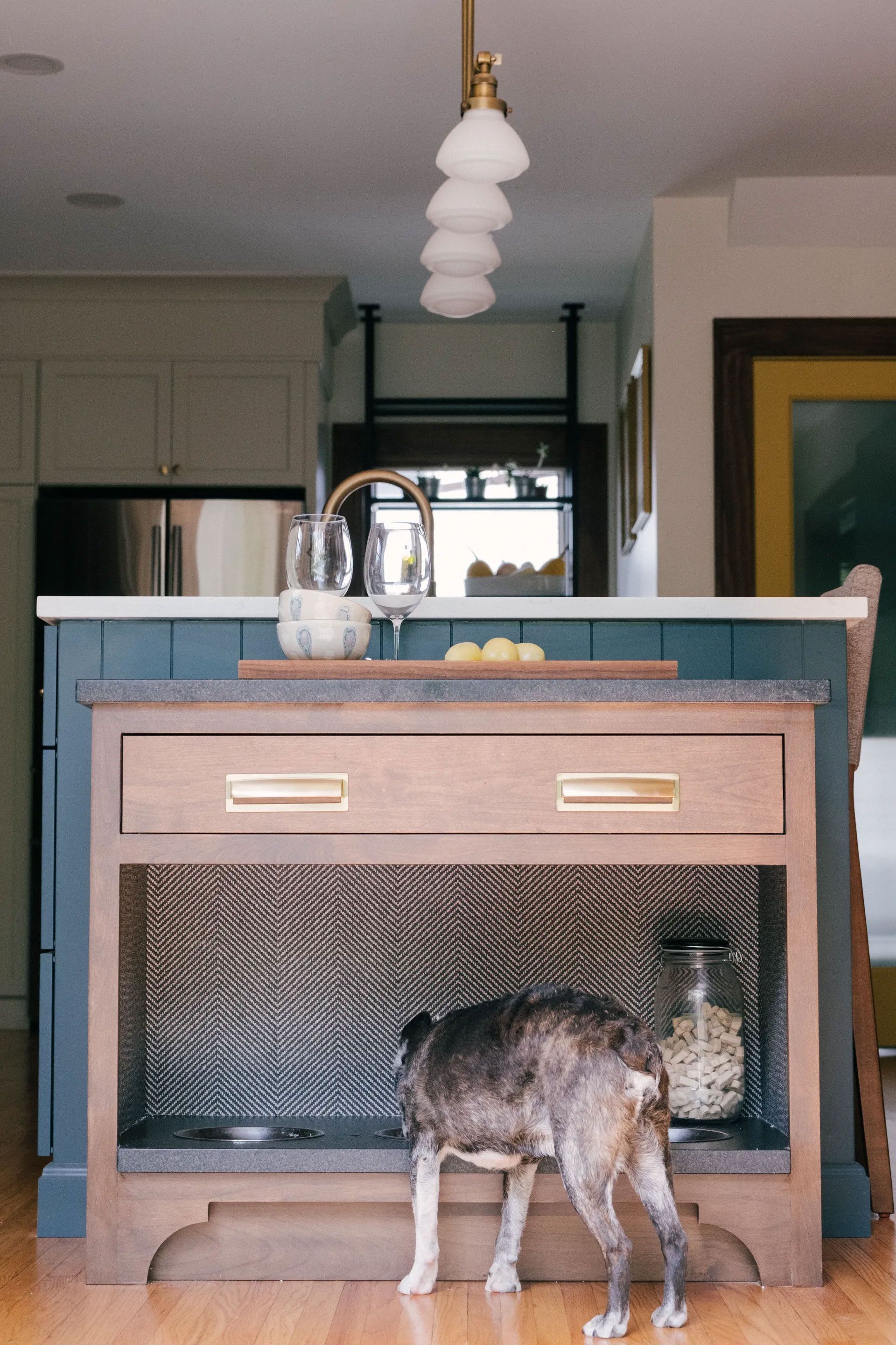 Dog entering built-in dog bed in a kitchen island. The island has drawers, a countertop, and decorative metal front.