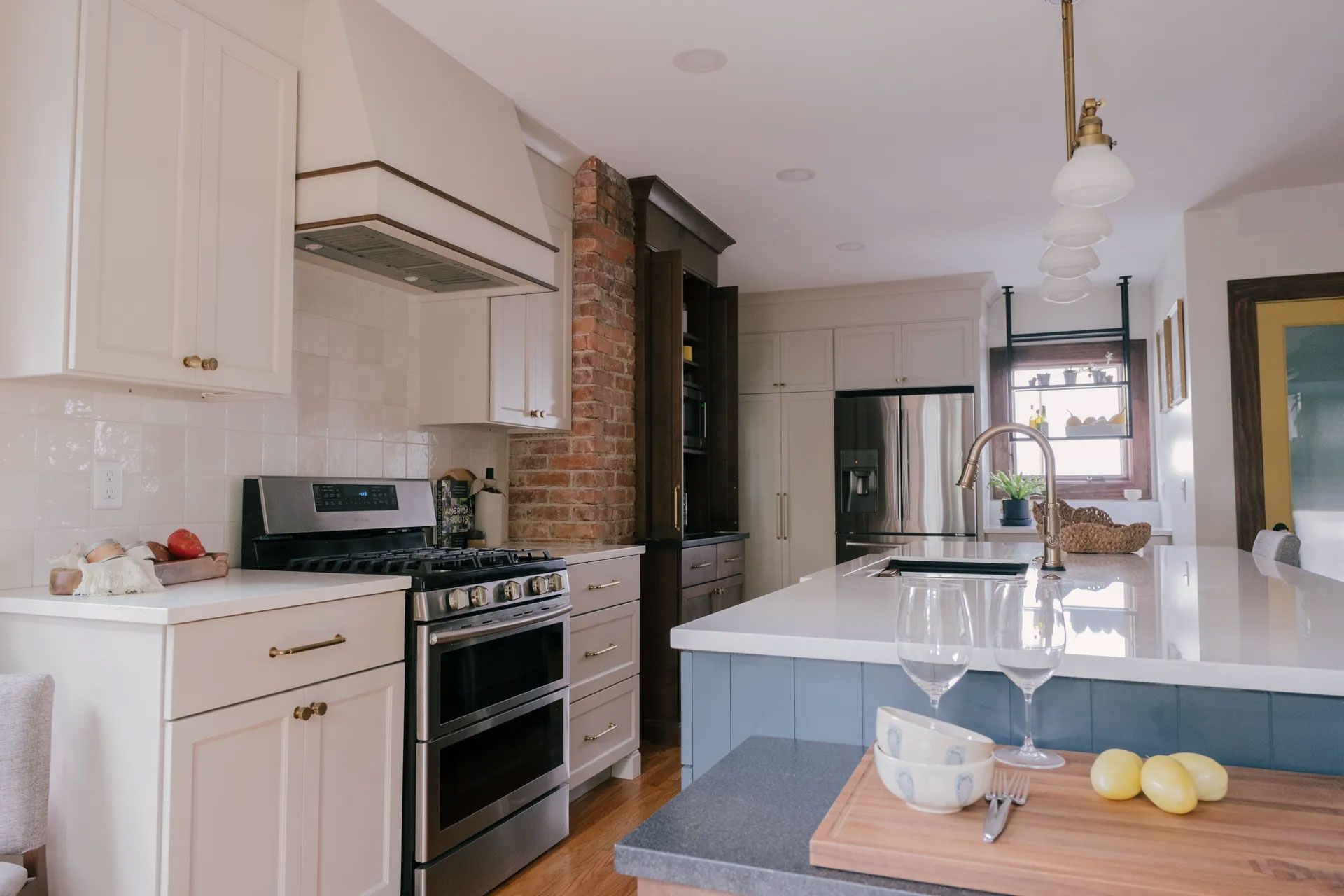 Kitchen with white cabinets, stainless steel appliances, blue island, exposed brick, and brass fixtures.