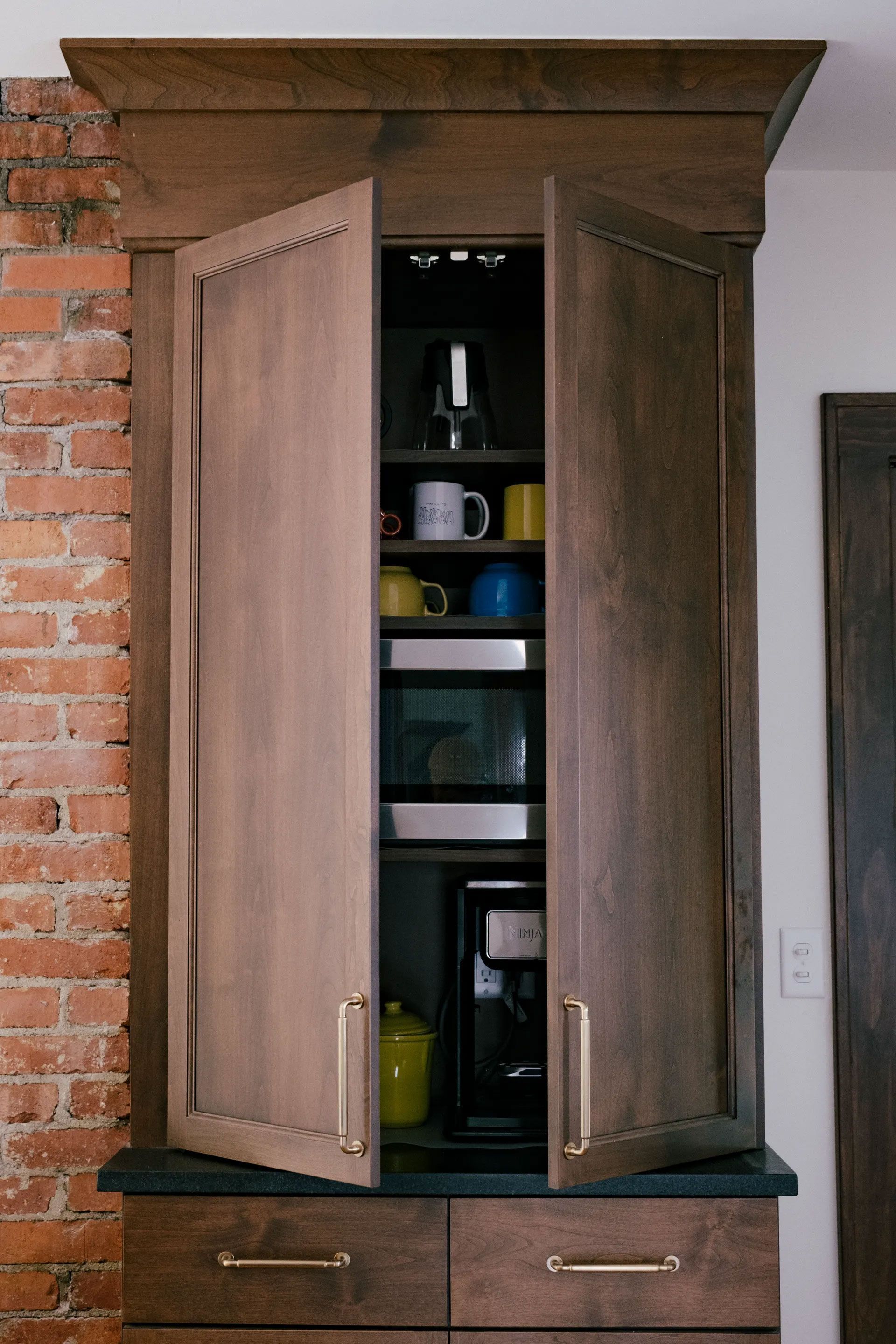 Built-in coffee cabinet with open doors, housing a microwave, coffee maker, and mugs, against a brick wall.