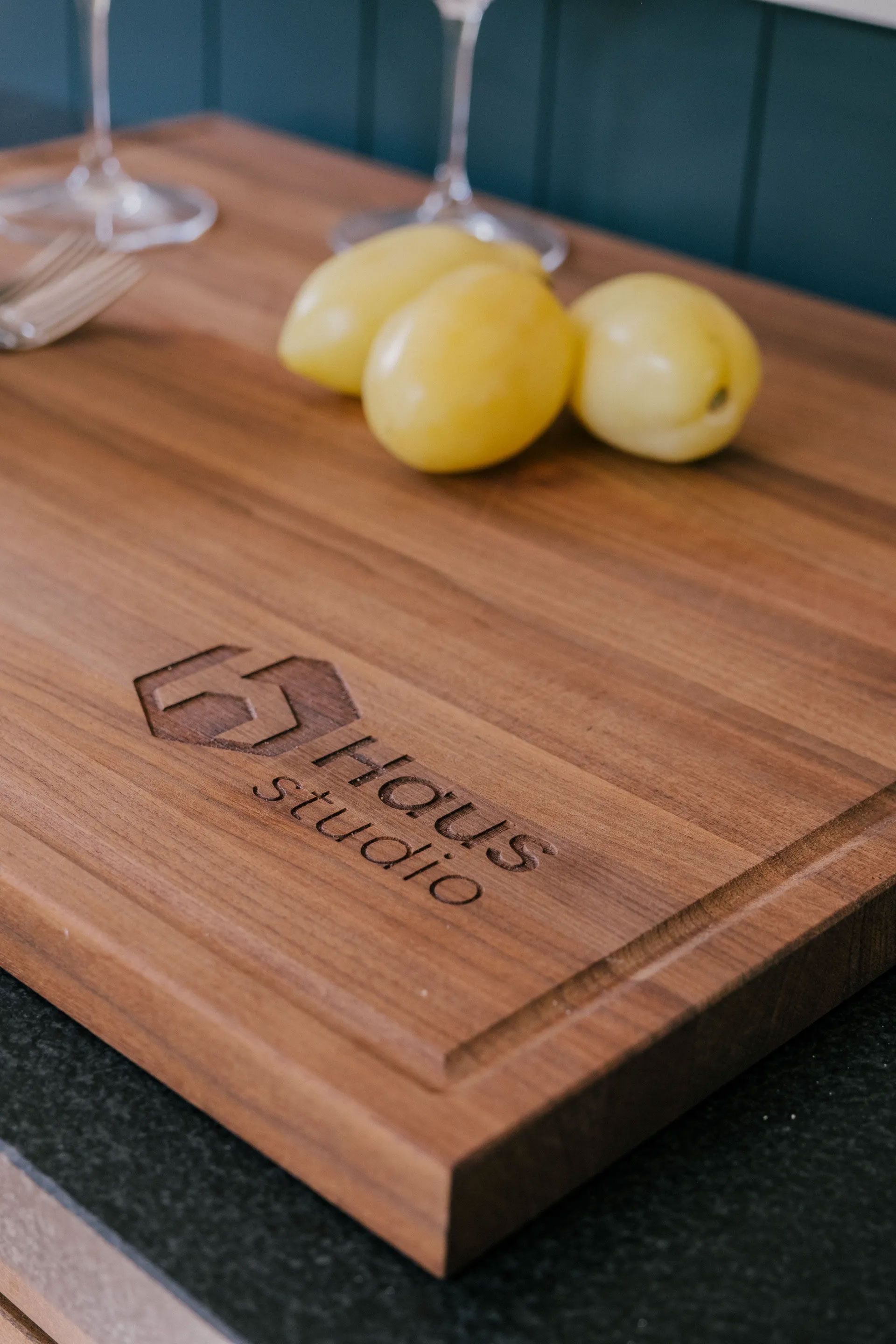 Wooden cutting board with lemons and logo, near wine glasses.