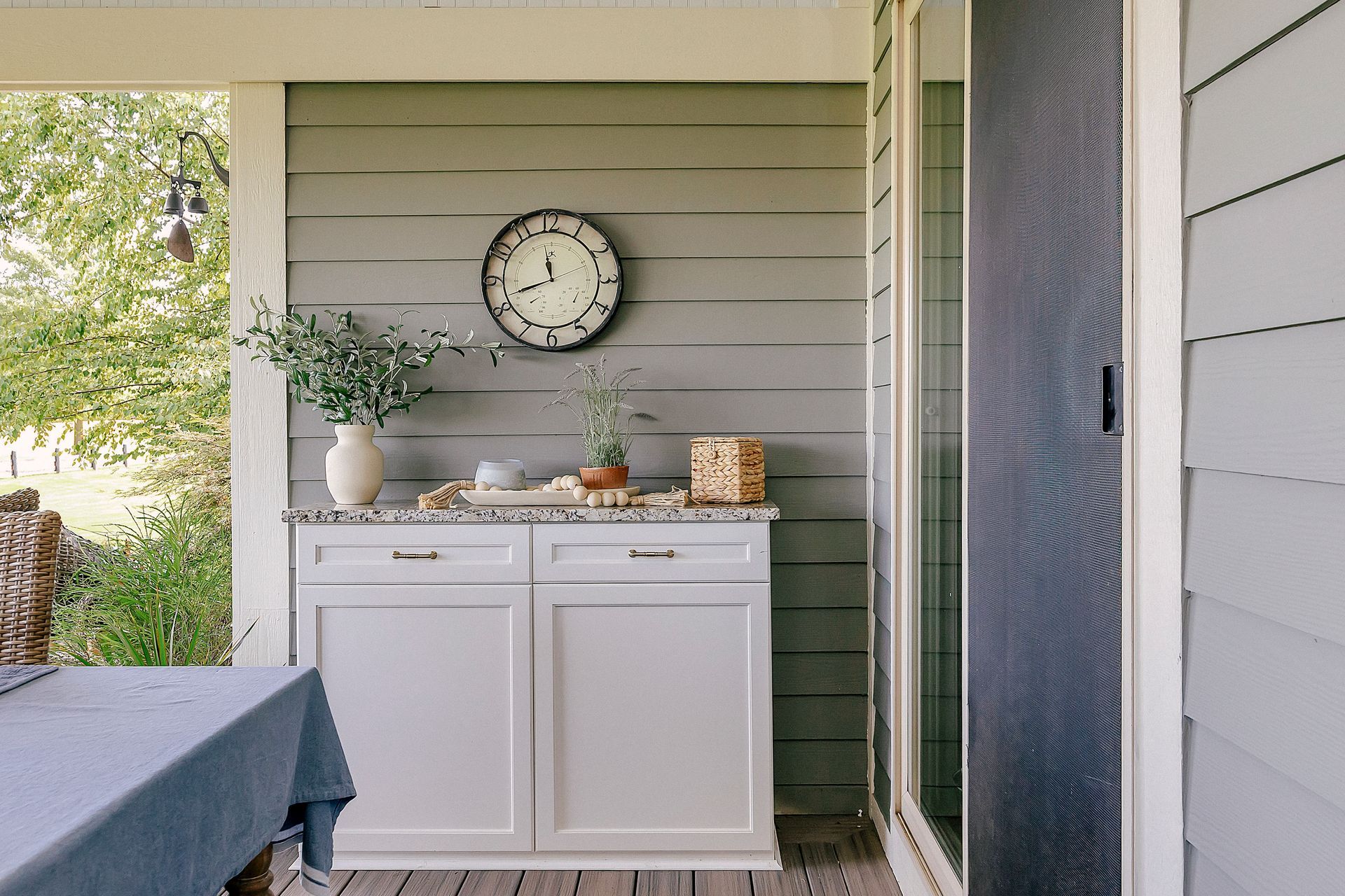 Outdoor kitchen area with gray cabinets, marble counter, clock, and greenery.