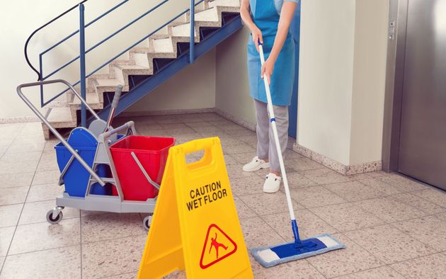 Custodian mopping a floor near a staircase and elevator, with a "Caution Wet Floor" sign and cleaning cart.