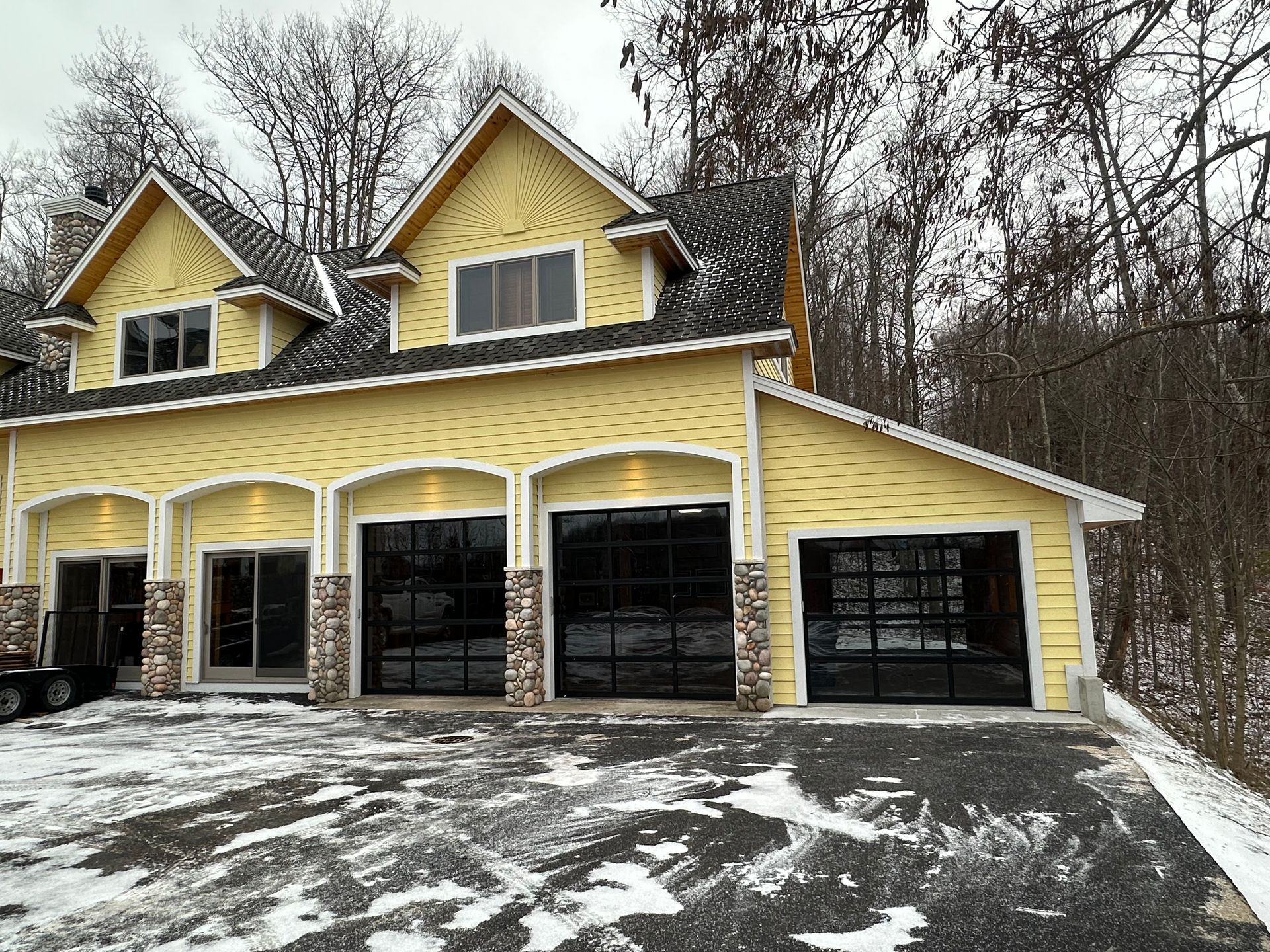 Yellow house with black garage doors, snowy driveway, and wooded background.