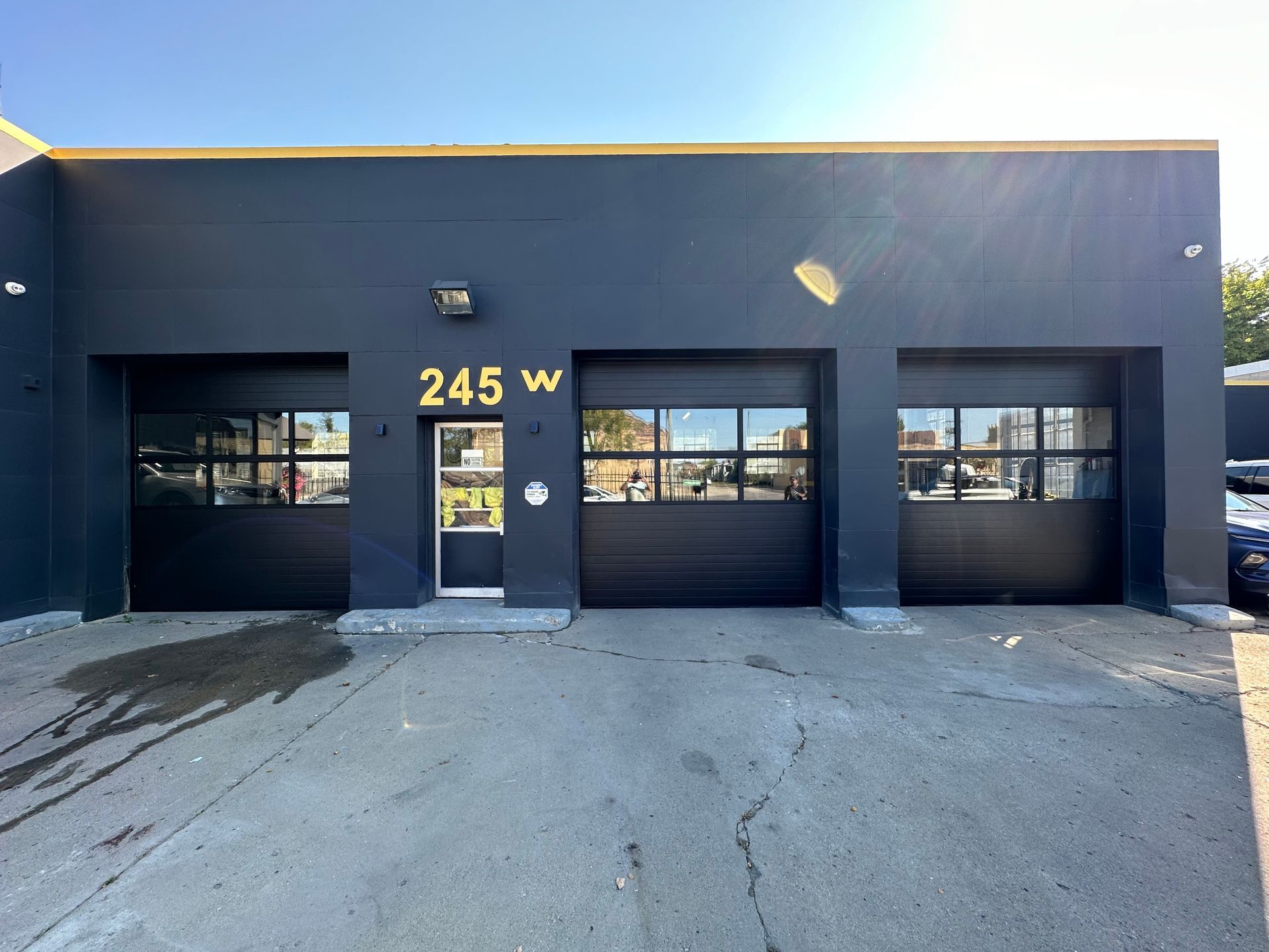 Dark gray building with three garage doors and a glass door; address 245 W. Sunny day.