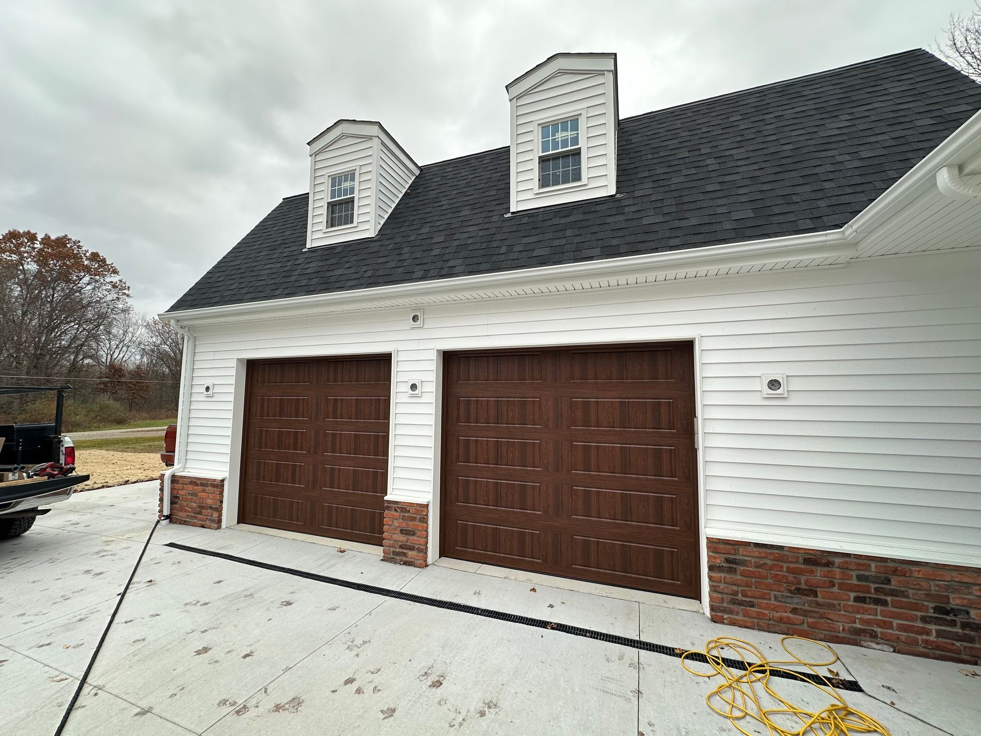 Two-car garage with brown doors, white siding, black roof, and brick accents. Dormers on the roof.
