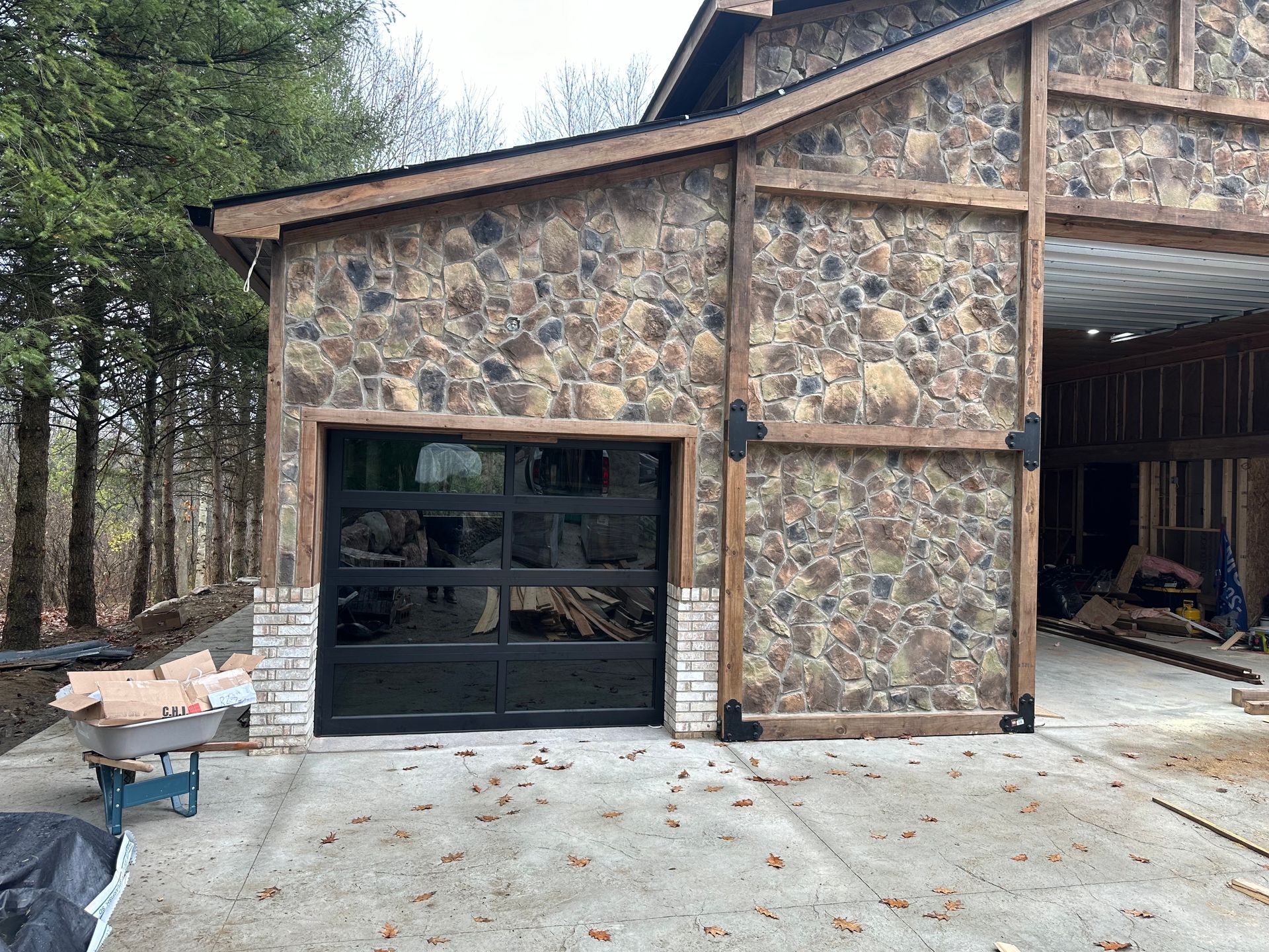 Stone-walled building with black glass garage door and wooden accents. Exterior shot with concrete driveway and trees.