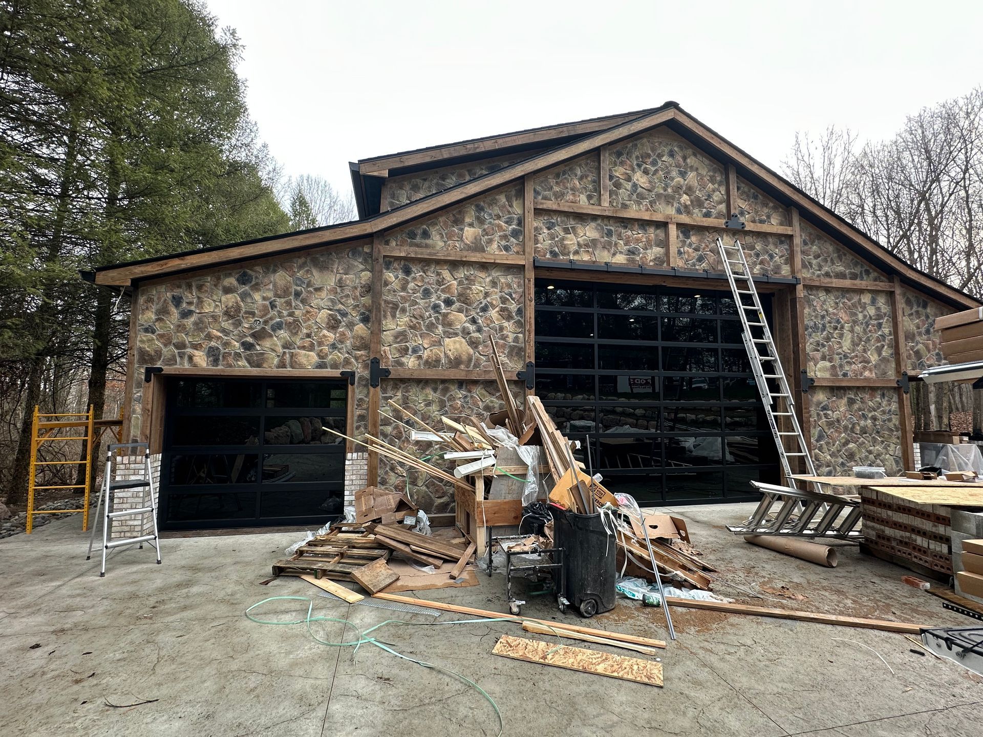 Barn-style building with stone siding and two black garage doors under construction, with a ladder and debris.