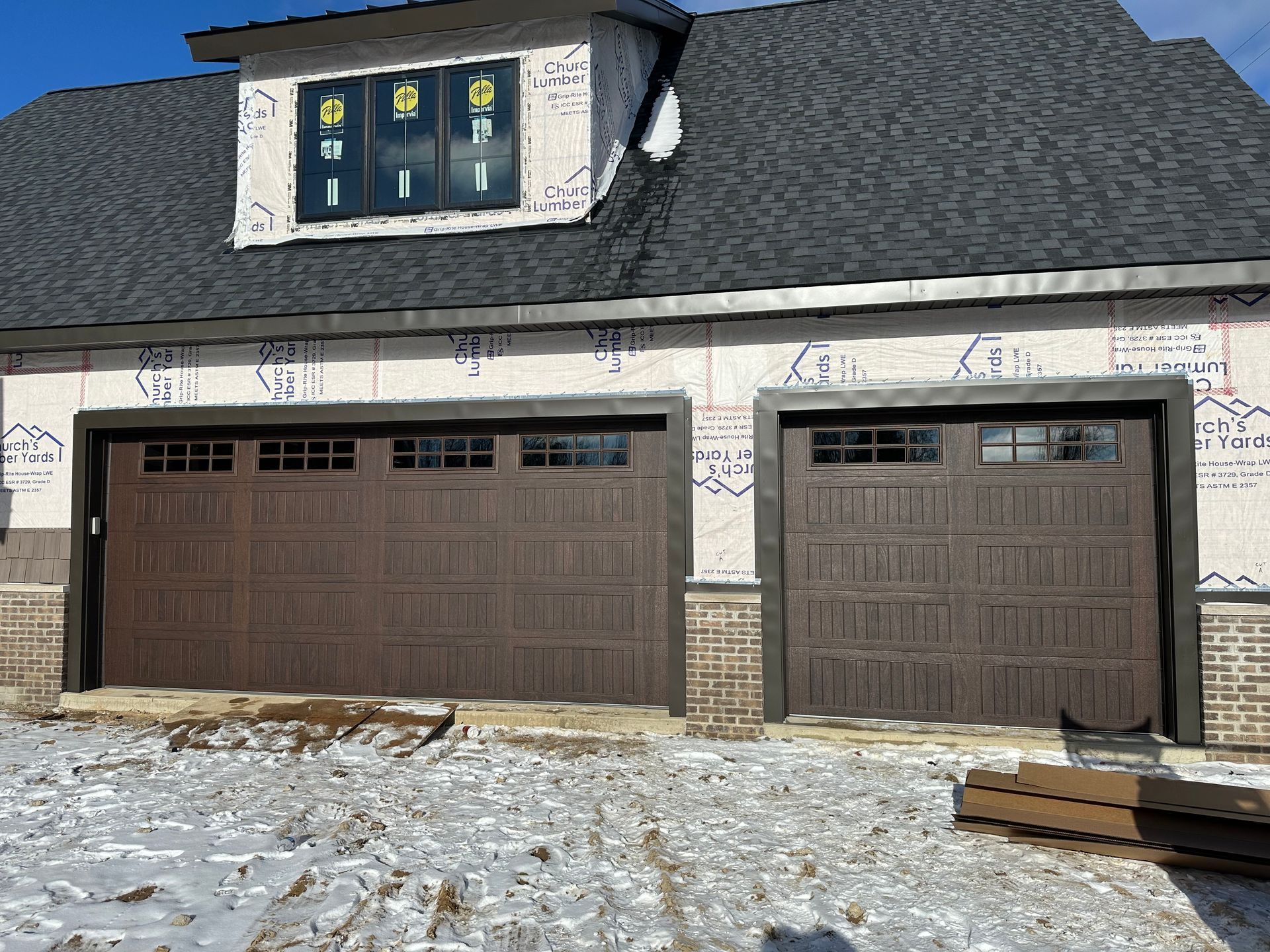 Two brown garage doors, dormer window, building under construction with snow on the ground.