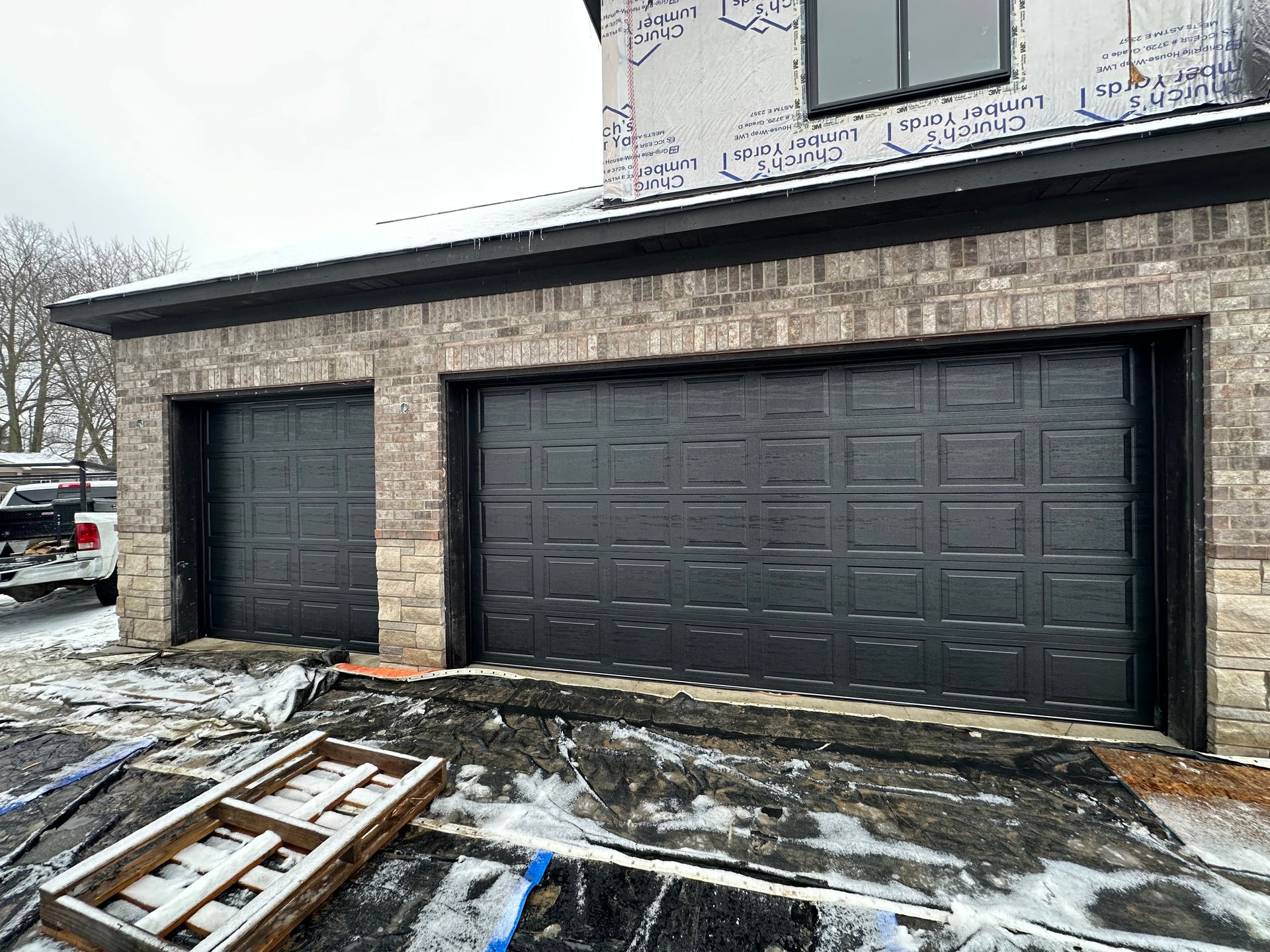 Two black garage doors on a brick building with snow on the ground and a wooden pallet.