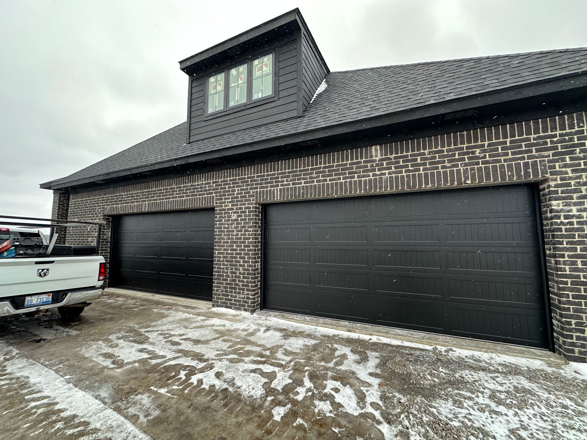 Black garage doors on a brick building with a dark roof and a small attic-like structure. Snow on the ground.