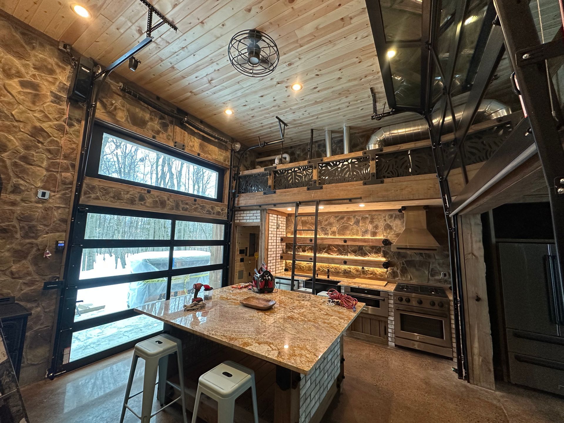 Kitchen with stone walls, island, stove, and a glass garage door overlooking snowy trees.