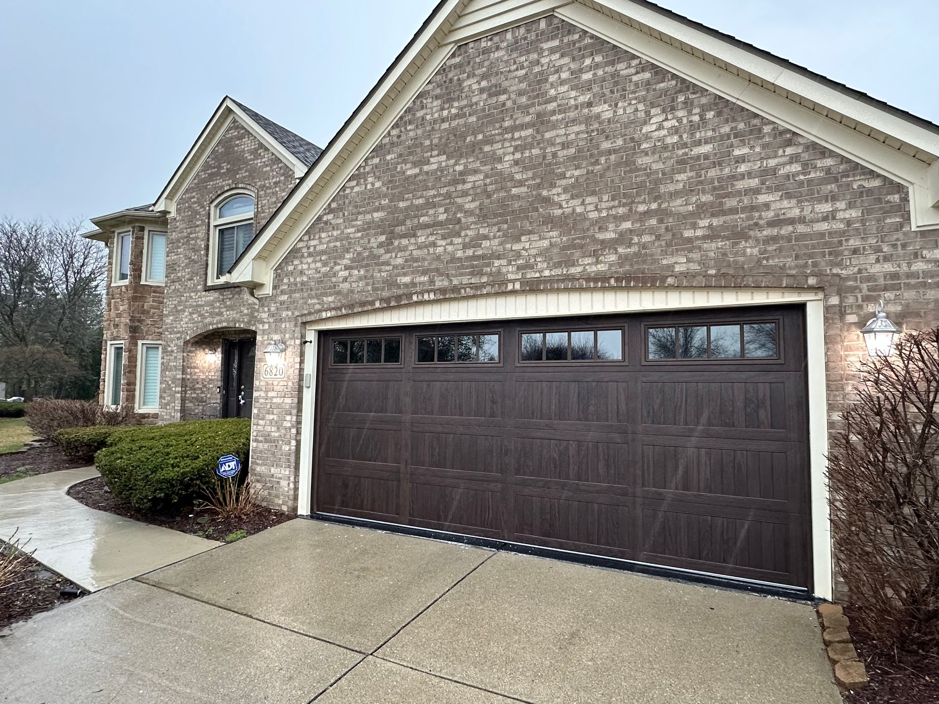 Brown garage door on a brick house with a wet driveway on an overcast day.