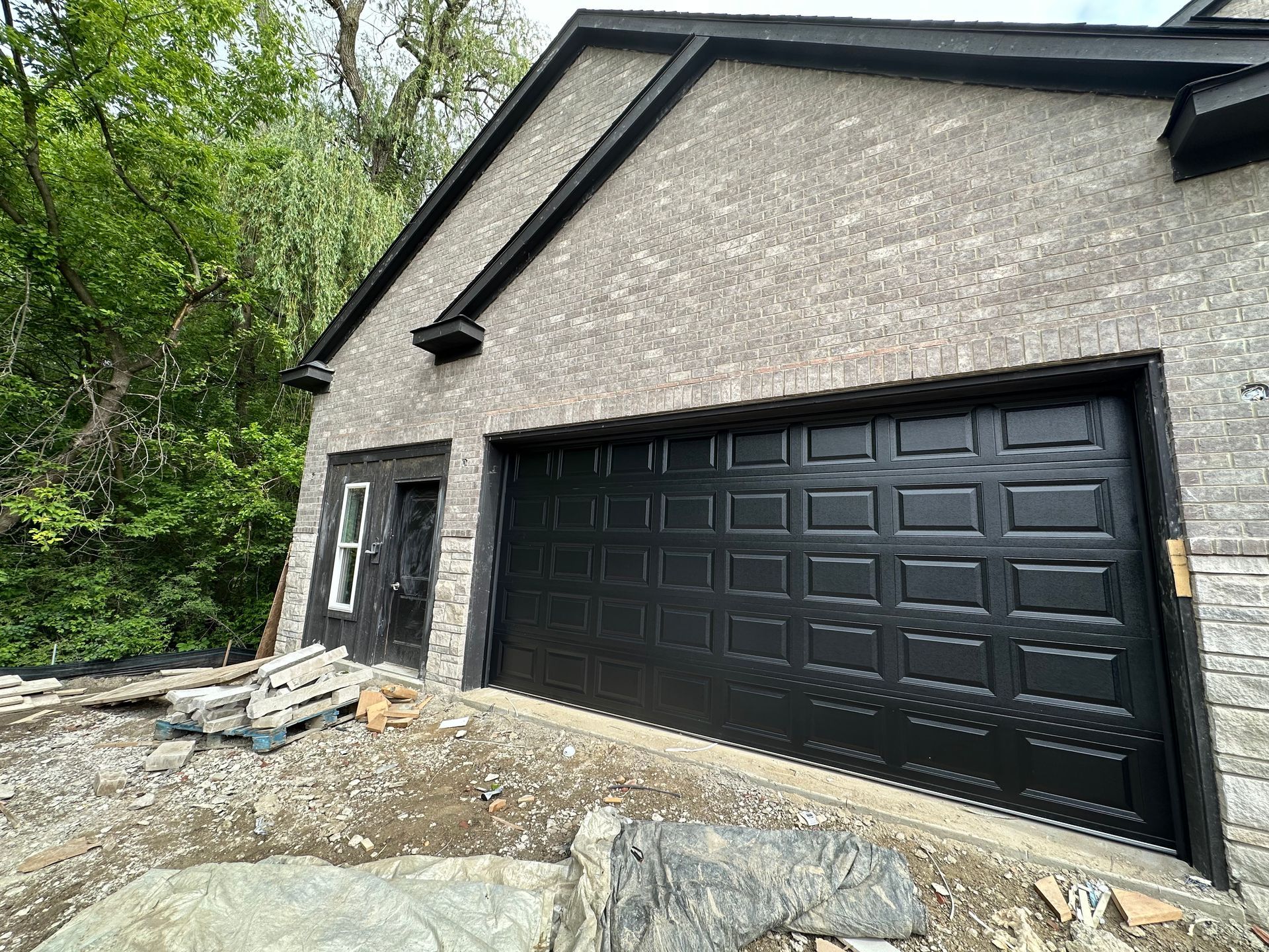 Black garage door on a brick building with a small door to the side; set outdoors, under a dark roof.