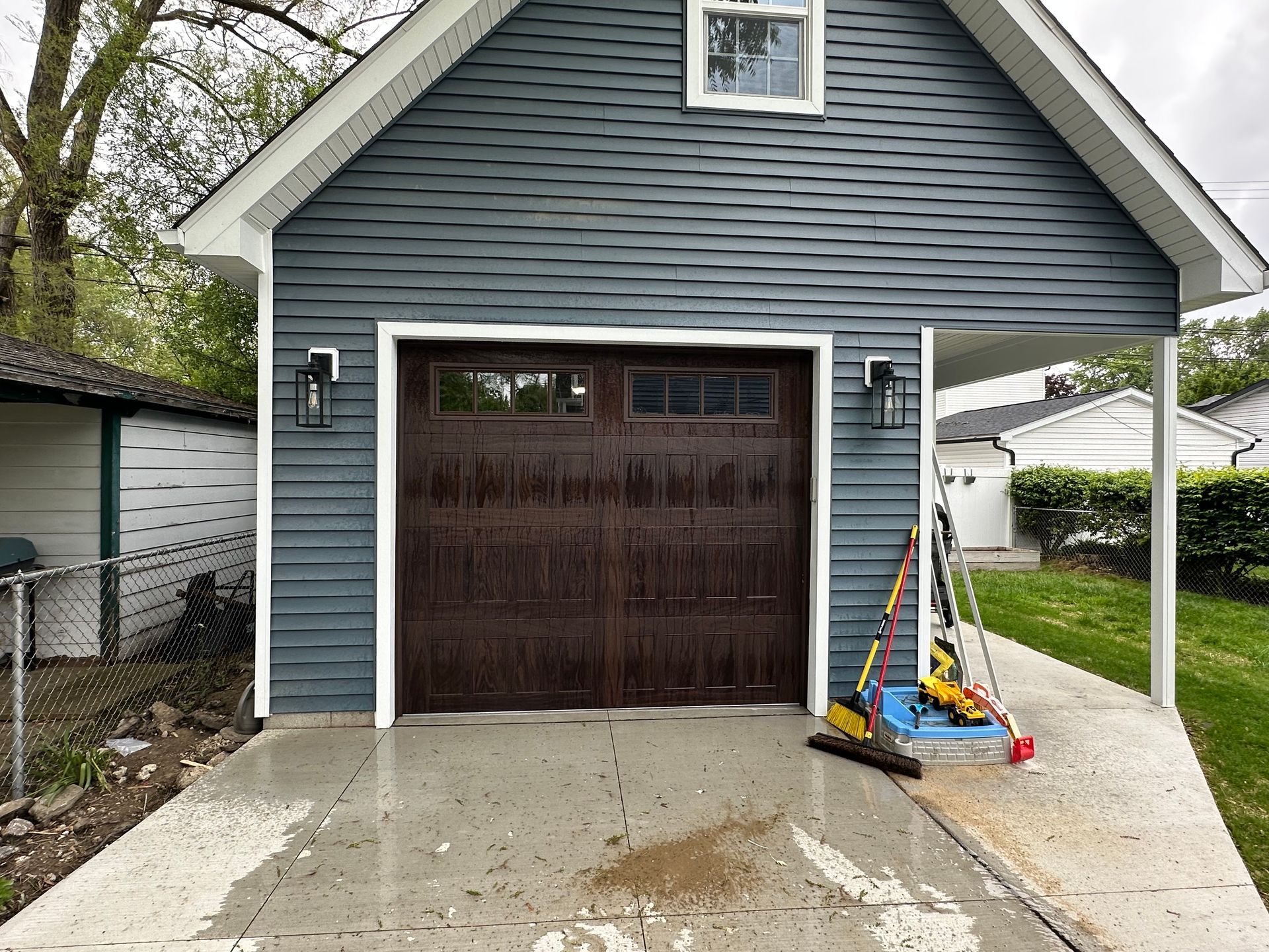 Dark brown garage door, blue siding, white trim on a detached garage. Tools lean against the side.