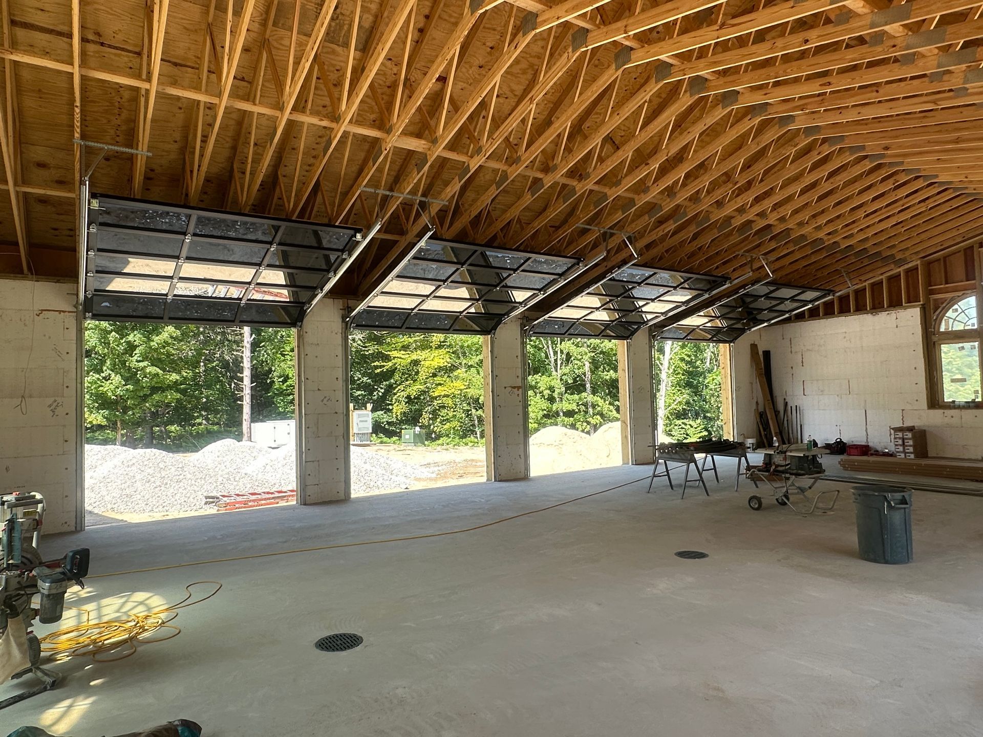 Interior of a building under construction, featuring open garage doors with glass panels, wooden beams, and concrete pillars.