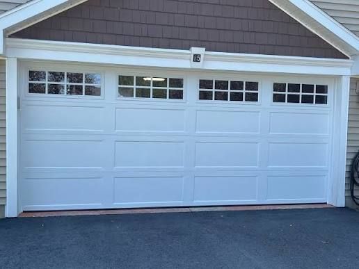 White garage door with rectangular windows, under brown roof, on asphalt driveway.