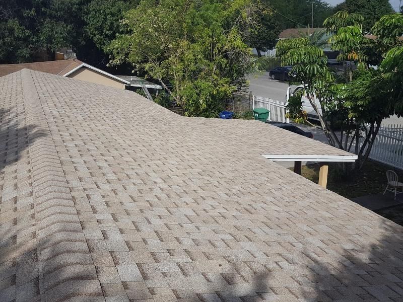 Tan asphalt shingle roof on a house, angled view with trees and a street in the background.