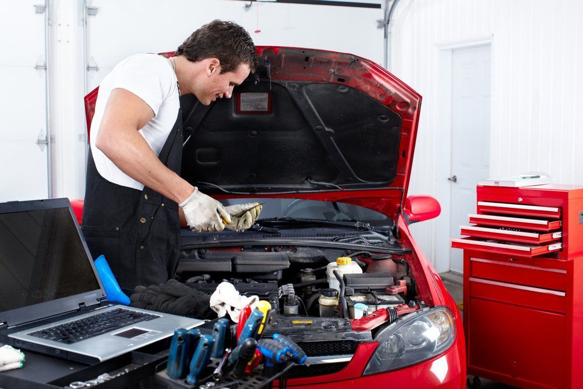 Mechanic inspecting red car engine, wearing overalls and gloves, with laptop and tools in garage.