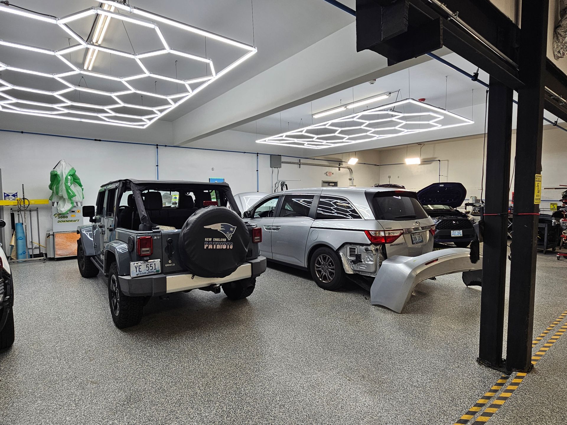 Jeep and SUV in a repair shop, lights overhead. A bumper lays on the floor.