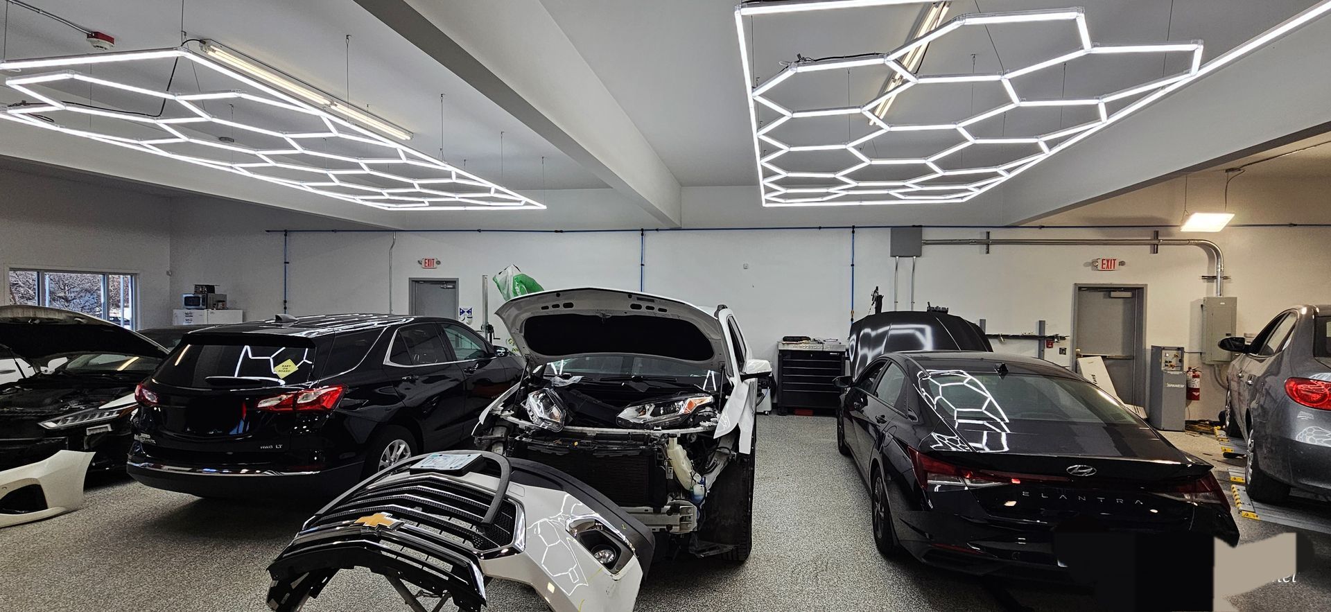 Cars in a workshop, under hexagonal lights, some with open hoods and removed parts.