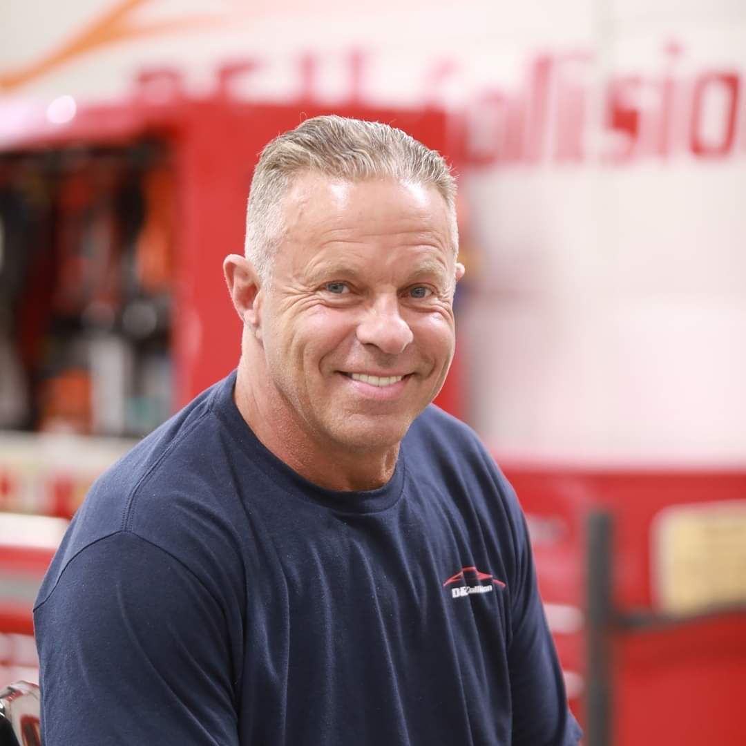 Man smiling, wearing a navy blue shirt, in front of a red and white building.