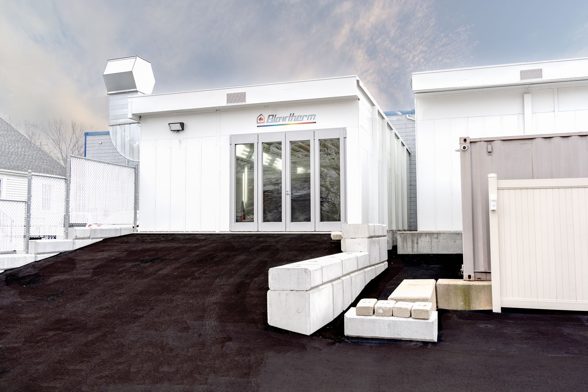 White building with glass doors, ramp, and concrete blocks. Black asphalt ground, cloudy sky.