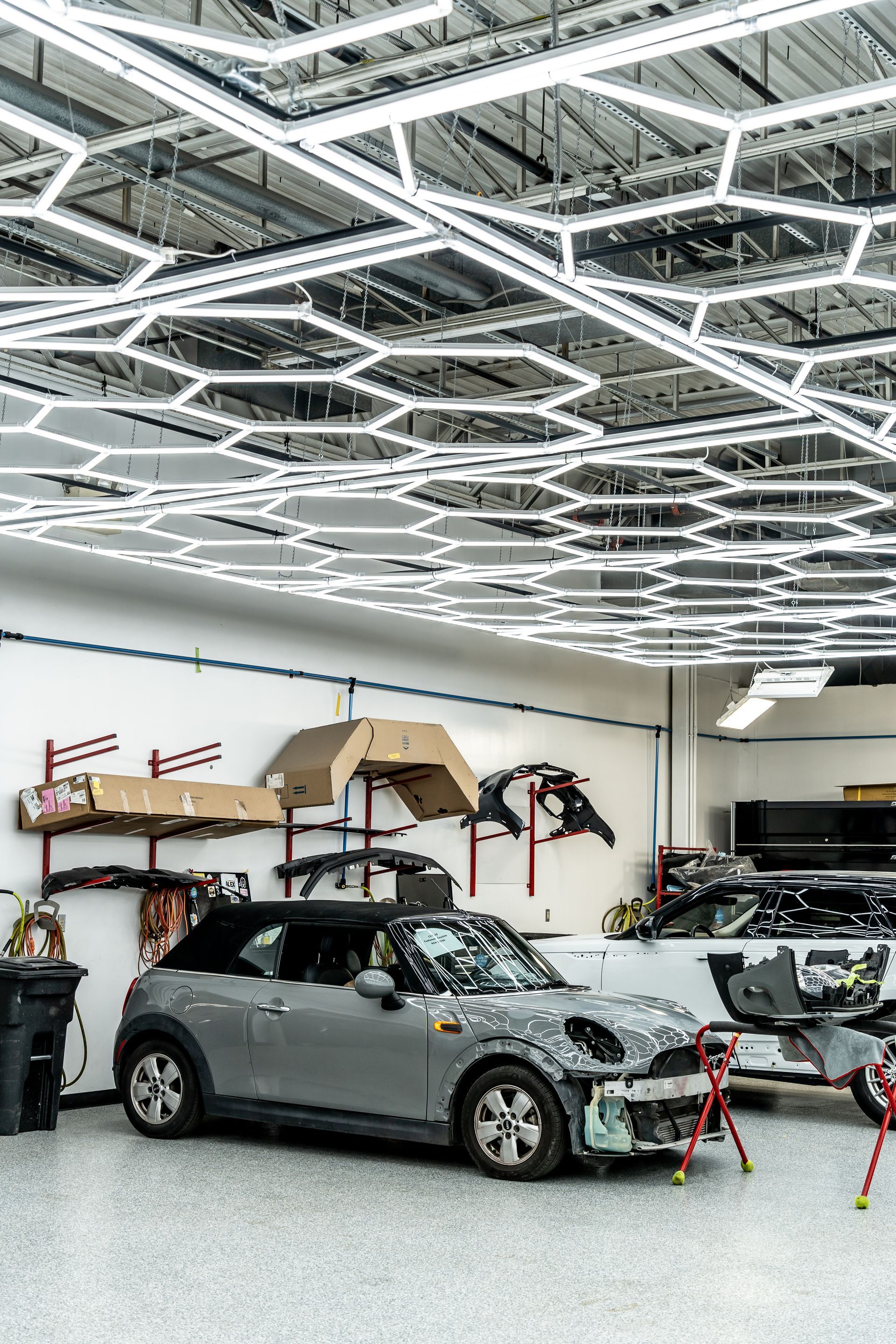 Grey Mini Cooper convertible in auto shop with hexagon-shaped overhead lights, parts and another car.