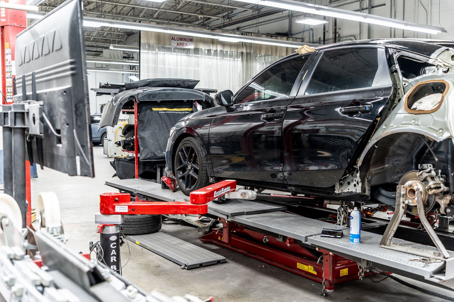 Black car on a lift in an auto body shop; two cars being repaired.