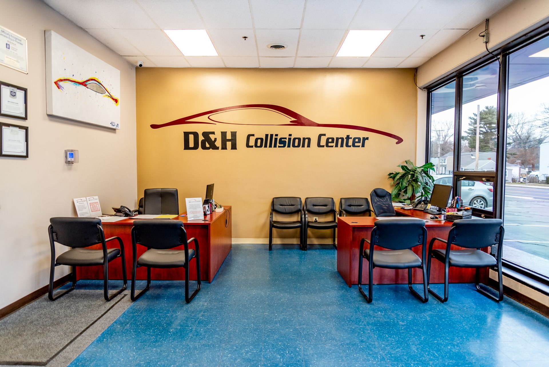Interior of D&H Collision Center. Reception area with desks, chairs, and logo on a tan wall. Blue flooring and large windows.