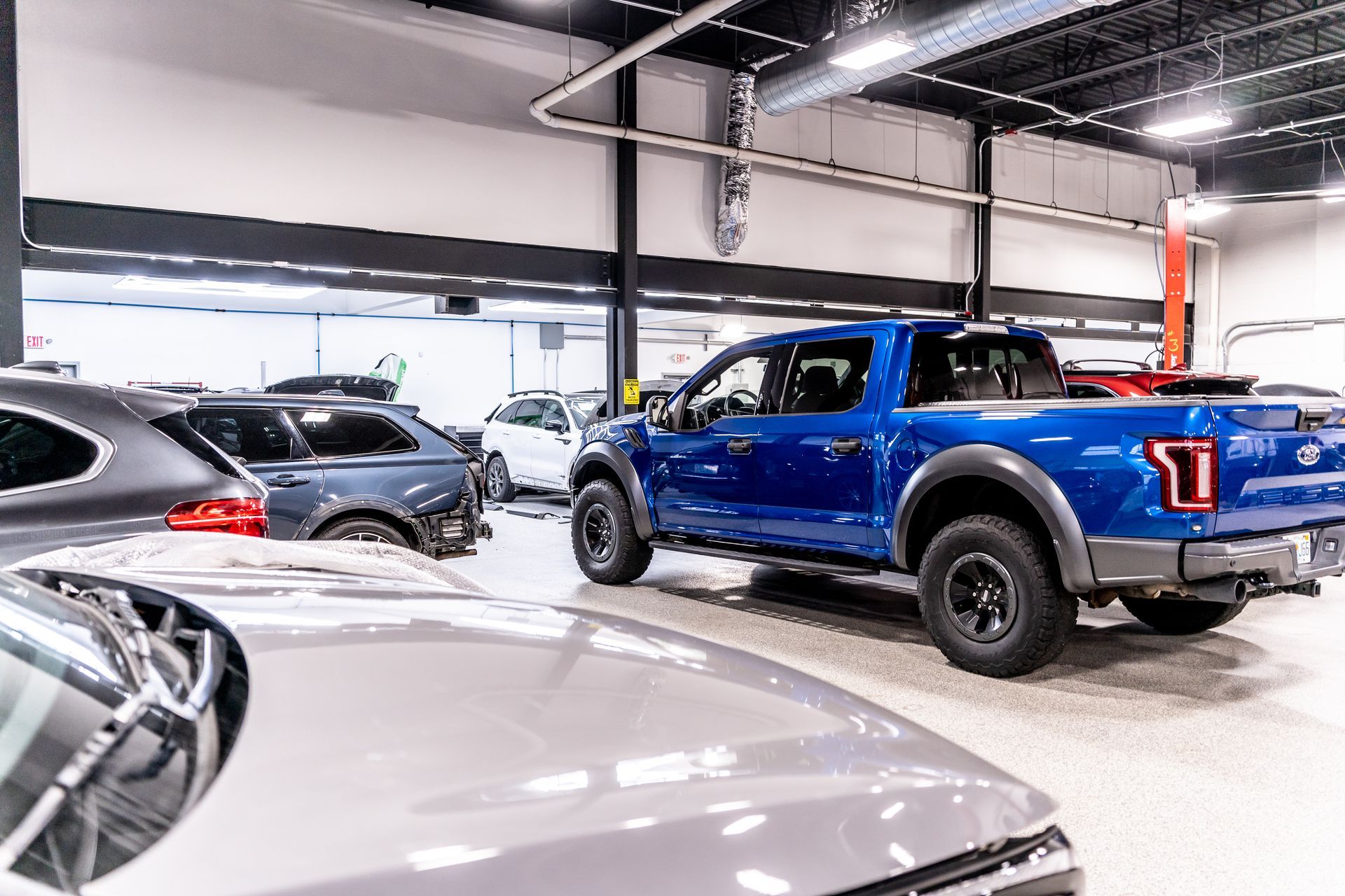 Blue Ford Raptor pickup truck inside a car showroom. Other vehicles are visible in the background.