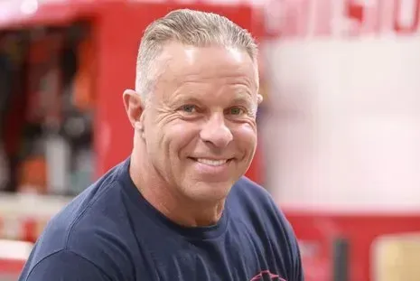 Man with short gray hair smiles in front of a red vehicle. He wears a navy blue shirt.
