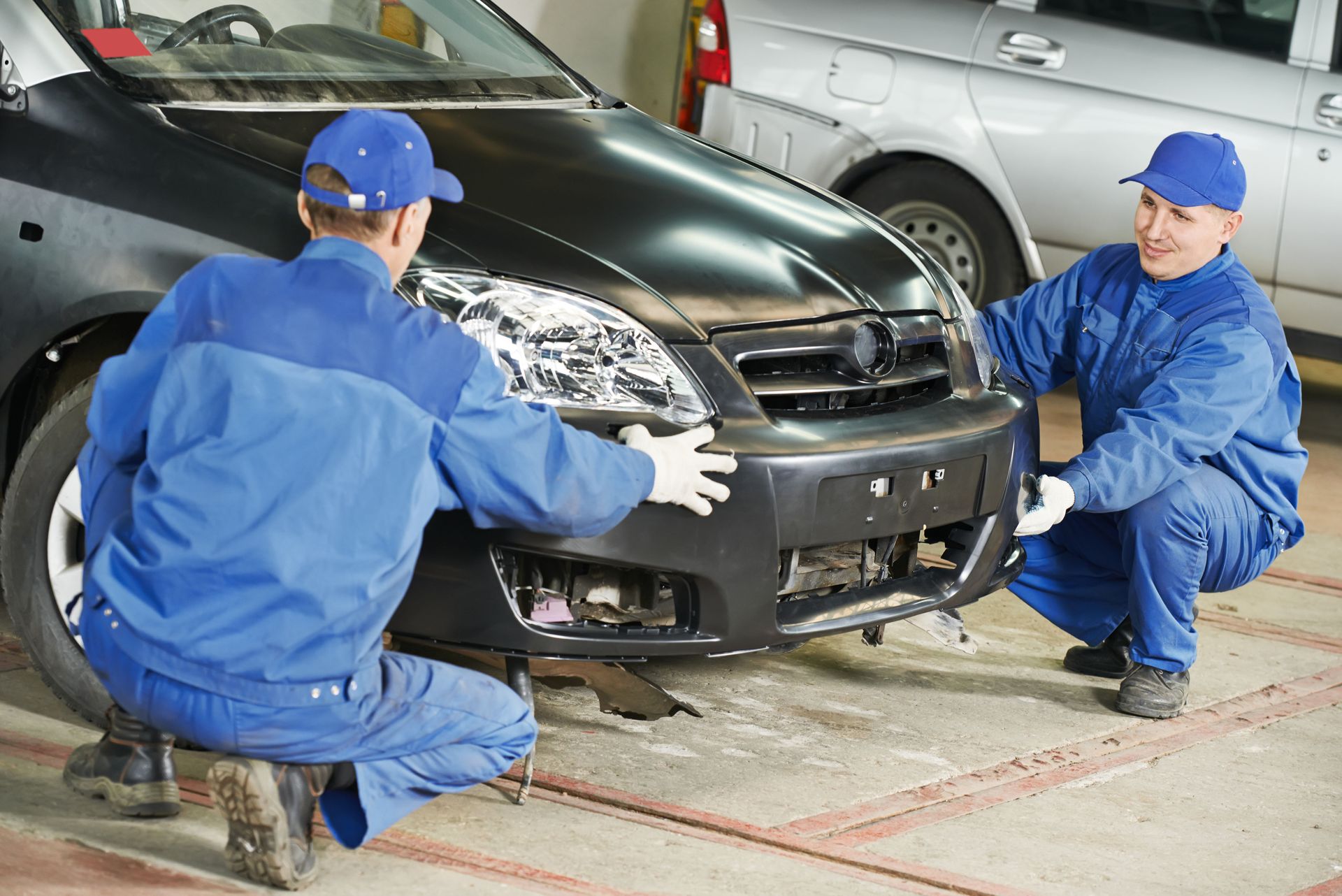 Two mechanics in blue coveralls installing a black car bumper in a garage.