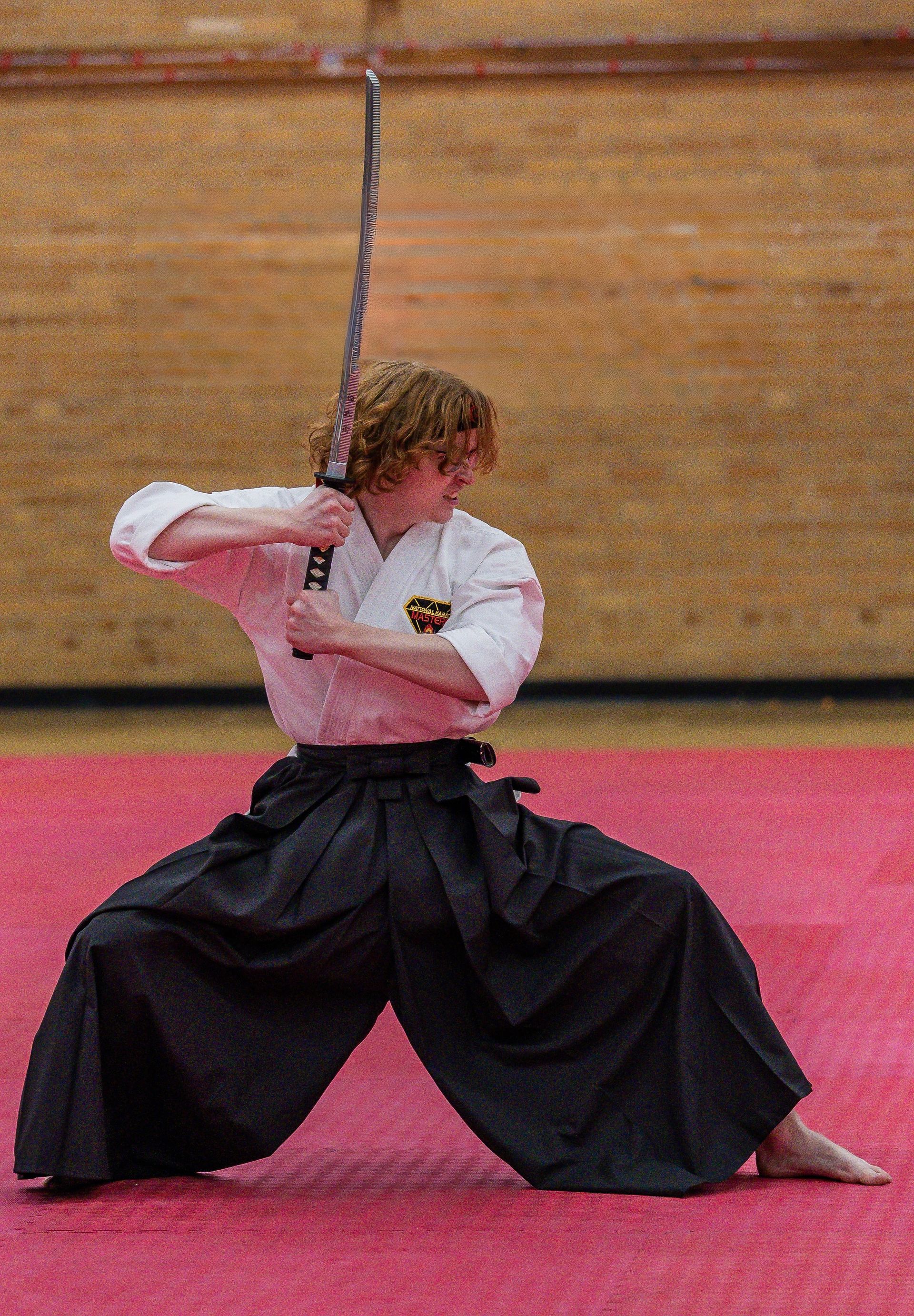 Martial artist in white uniform and black hakama posing with sword on pink mat indoors