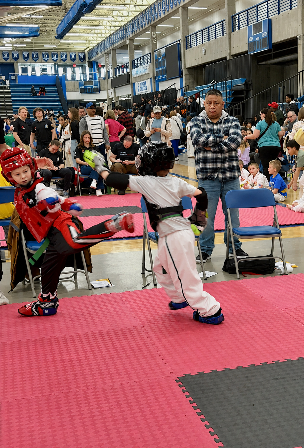 Two martial arts competitors spar on a red mat in an indoor event hall, with spectators watching nearby.