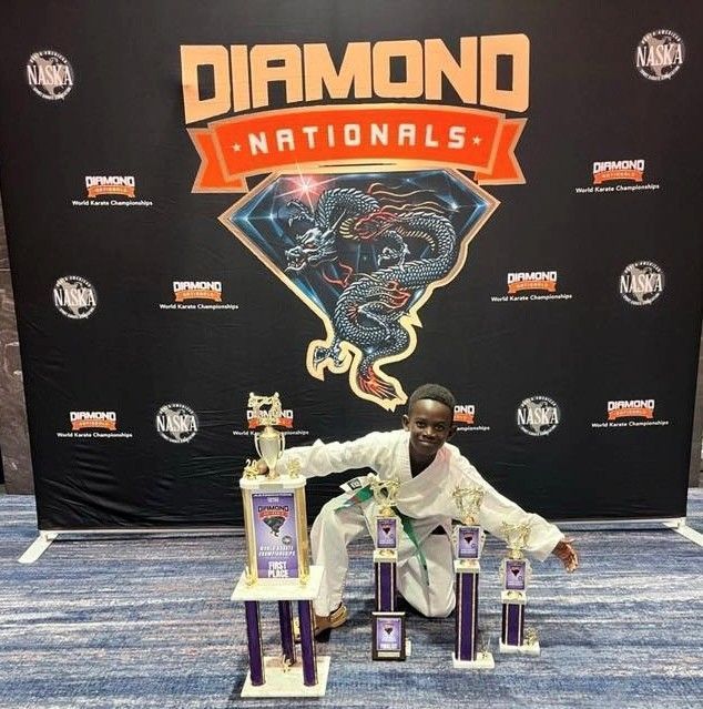 A karate athlete kneels in front of trophies at the Diamond Nationals event, wearing a white uniform and green belt.