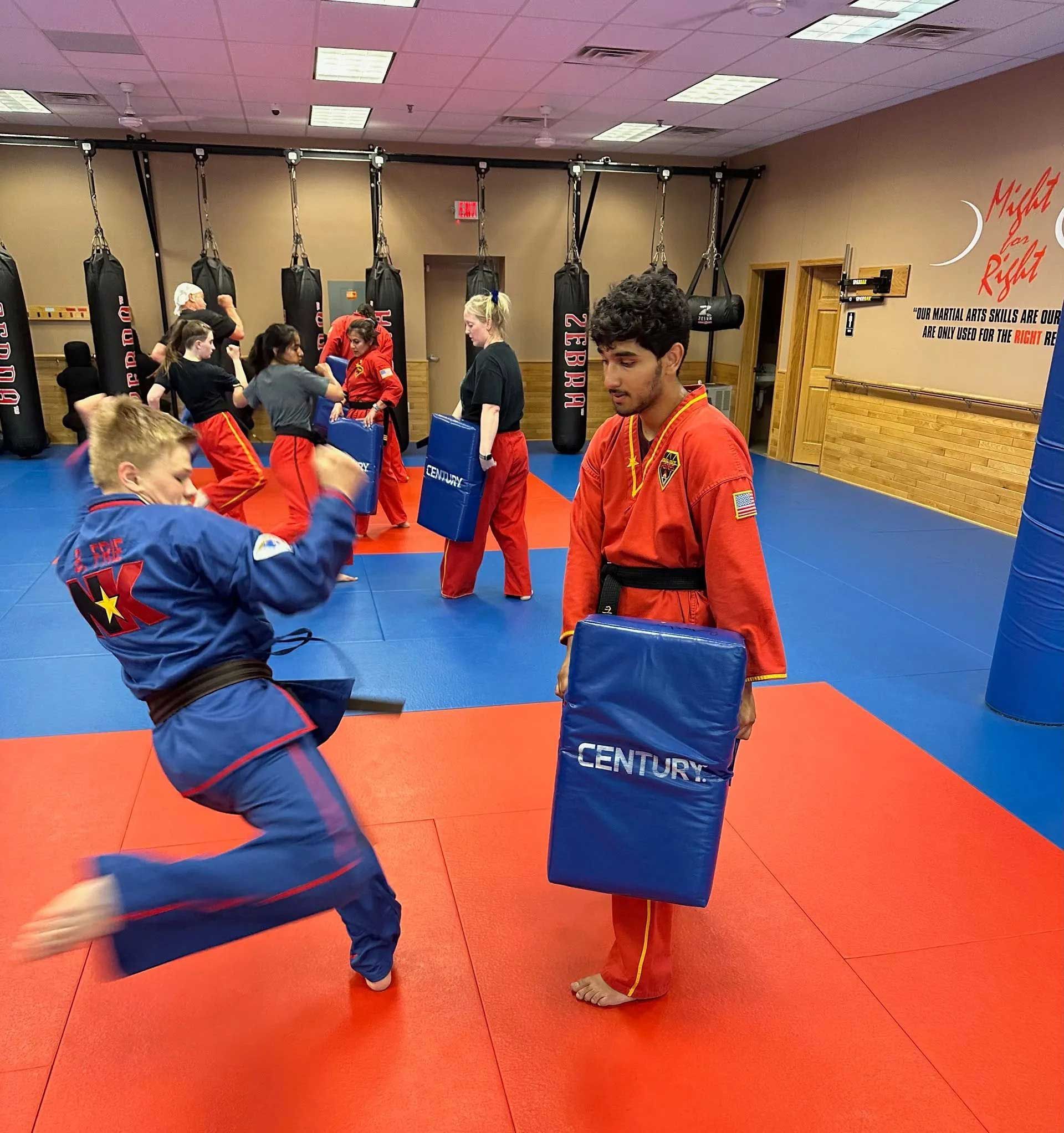 Martial arts class: Student in blue uniform kicks pad held by instructor in red uniform. Other students train.