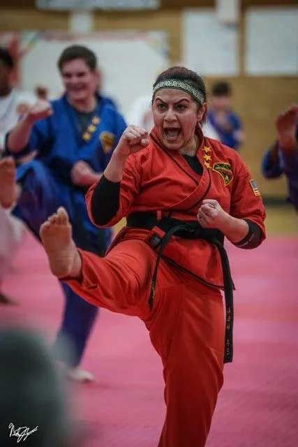 Woman in red karate uniform kicking, yelling, in a gym. Others in blue uniforms train in the background.