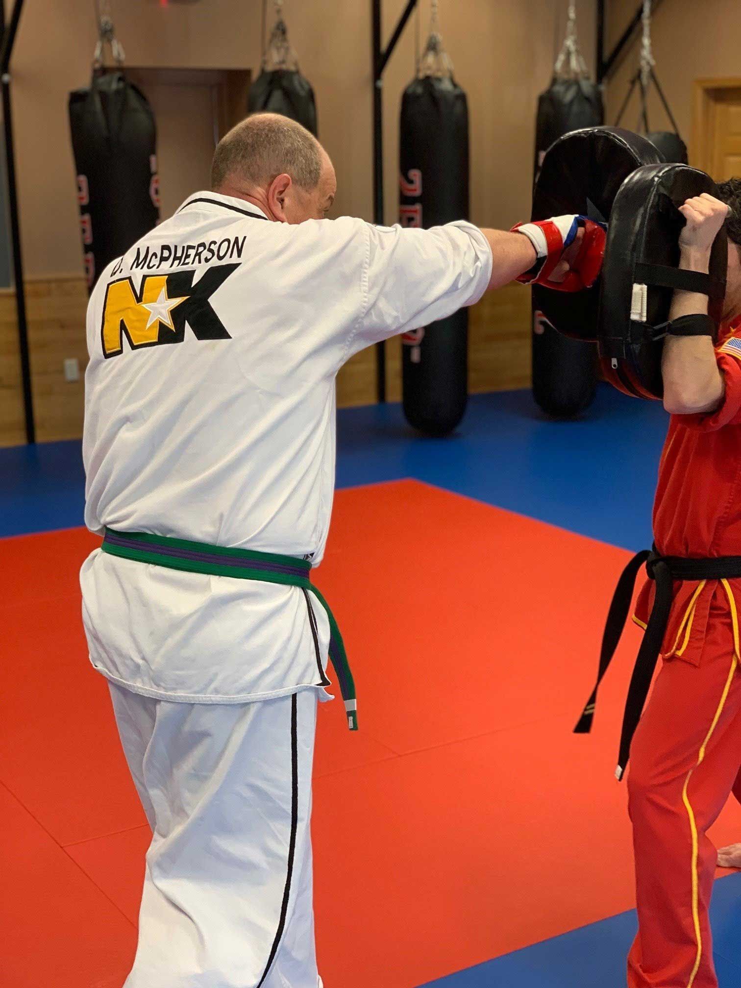 Martial artist in white uniform punches padded target held by person in red. Training in a gym.