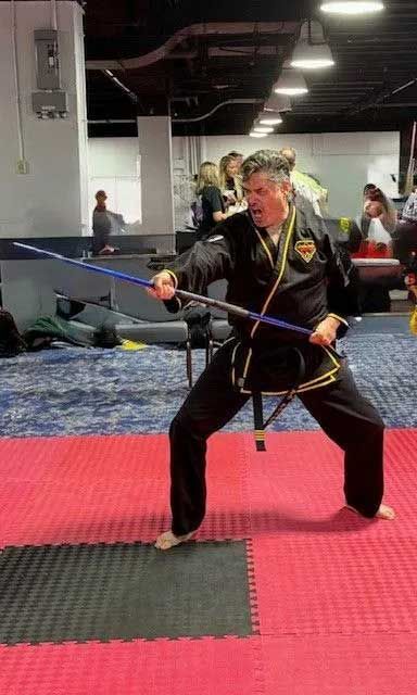 Martial artist in black uniform practices with blue bo staff on red mat. Indoor training space.