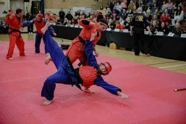 Martial arts competitors in red and blue uniforms sparring on a pink mat in front of spectators.