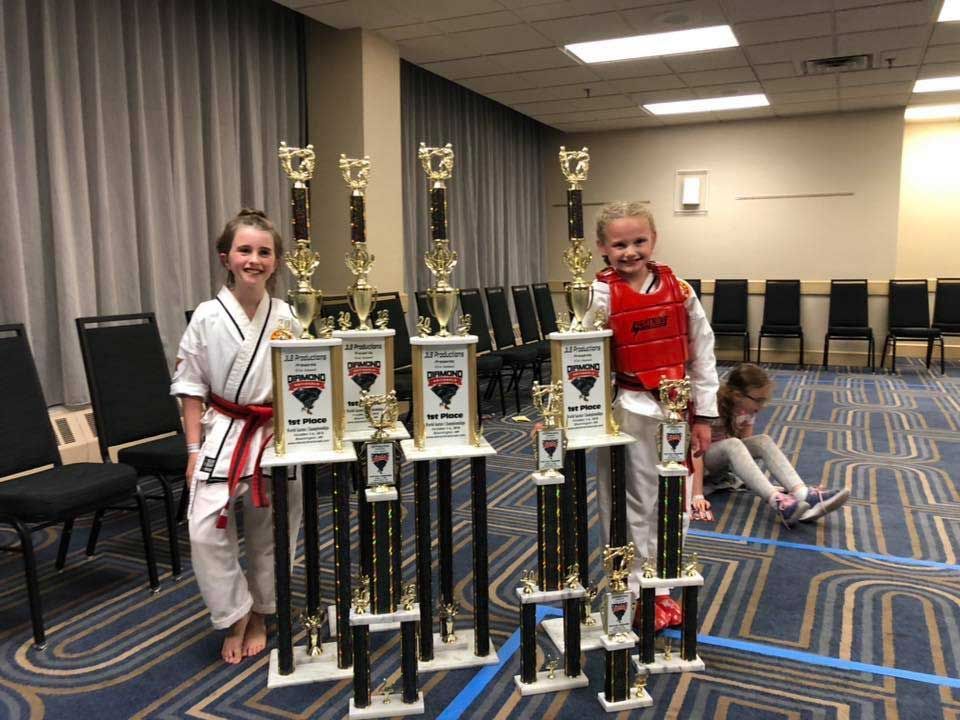 Two young girls in karate uniforms pose with trophies in a banquet hall.