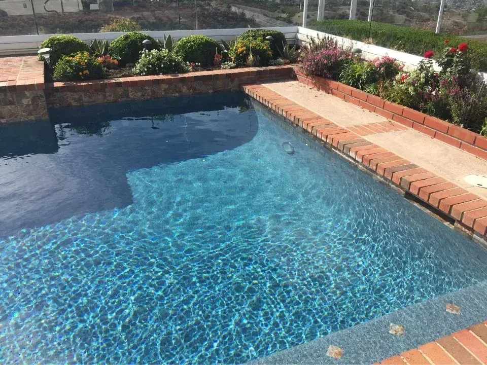 A rectangular swimming pool with brick trim and a pathway, surrounded by greenery and flowers.