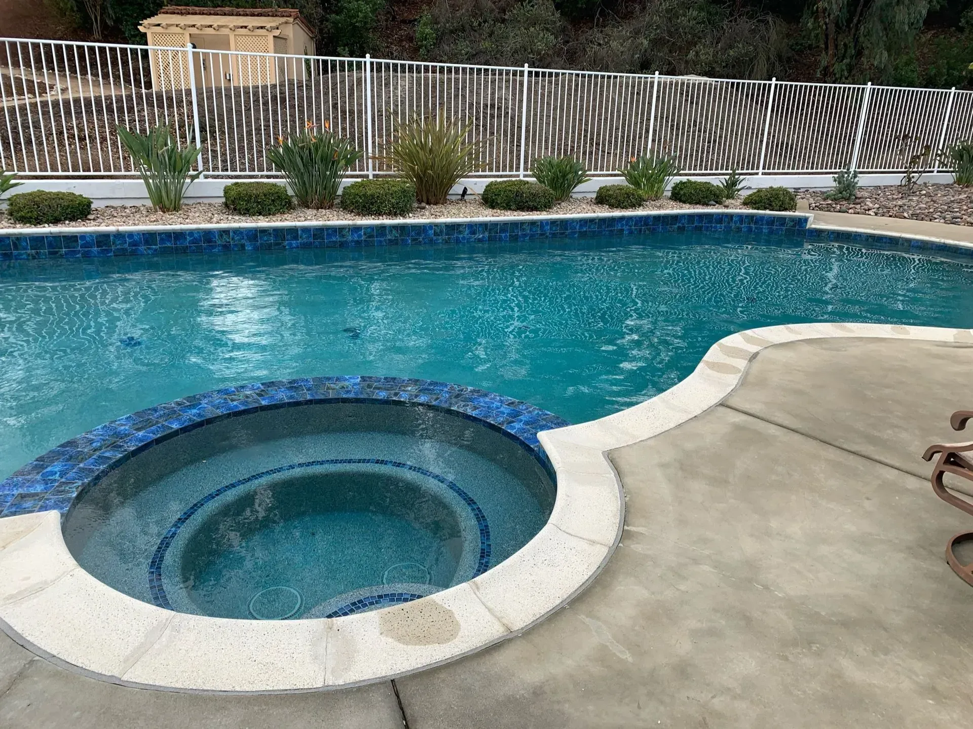 Pool with blue tile and hot tub, surrounded by concrete patio. A fence is in the background.