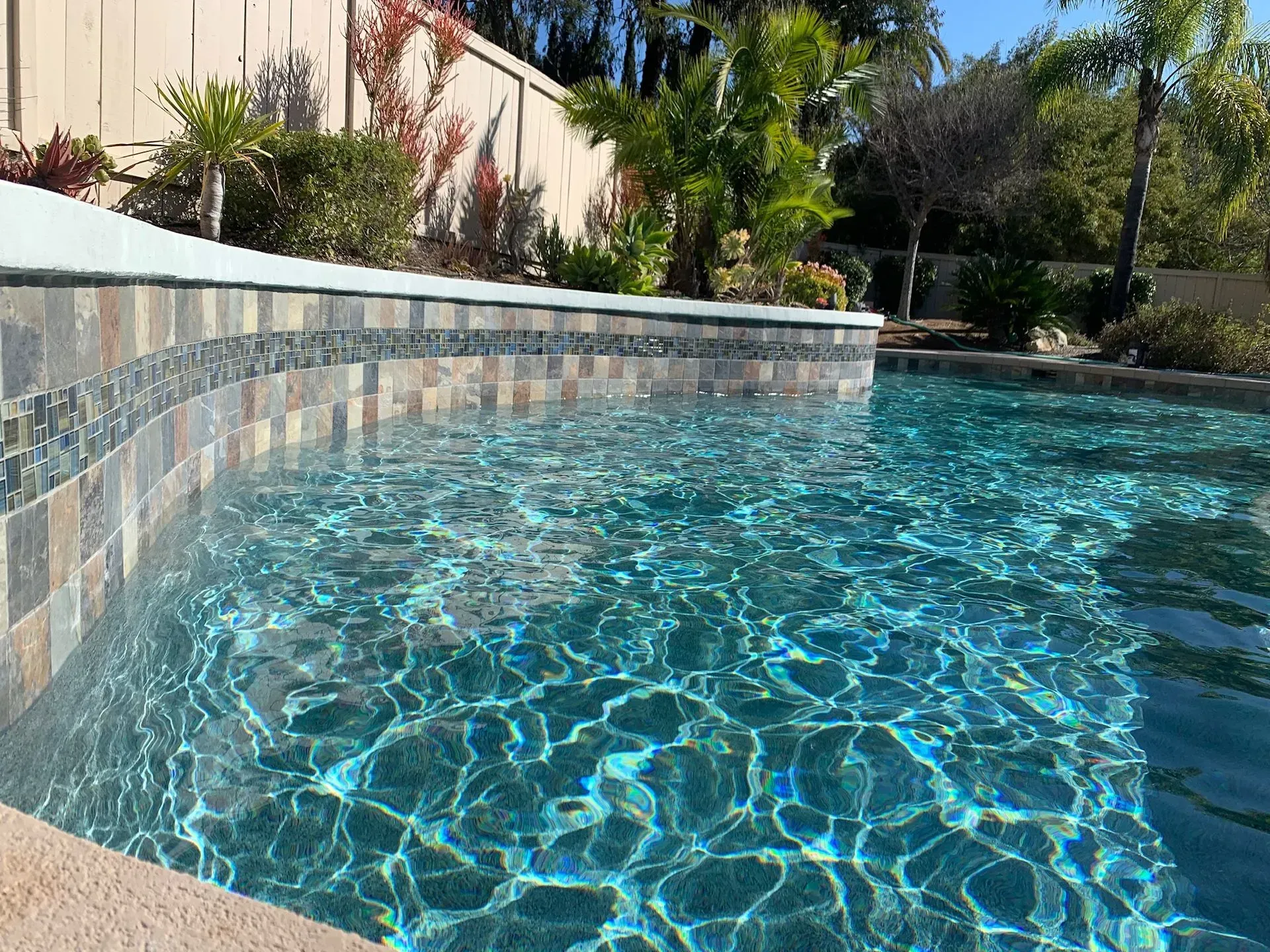 Pool with blue water and tiled edge, surrounded by greenery and a white fence.