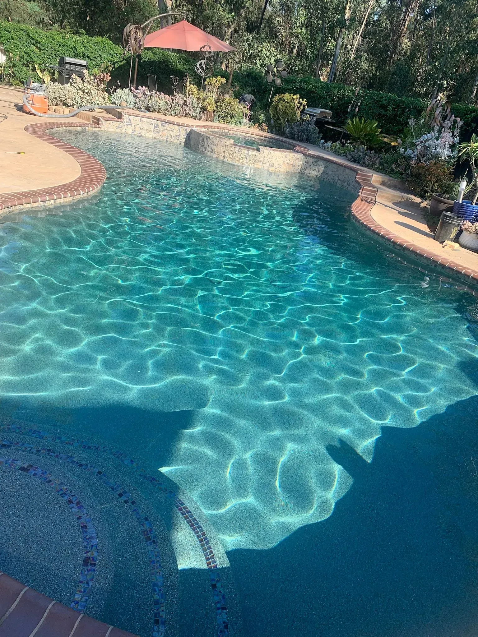 Swimming pool with turquoise water, surrounded by a stone deck and lush greenery. An umbrella shades a seating area.